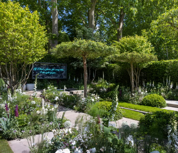 A lush garden with various trees, shrubs, and flowering plants, along with a pathway and a digital sign displaying a message in a tranquil outdoor setting.