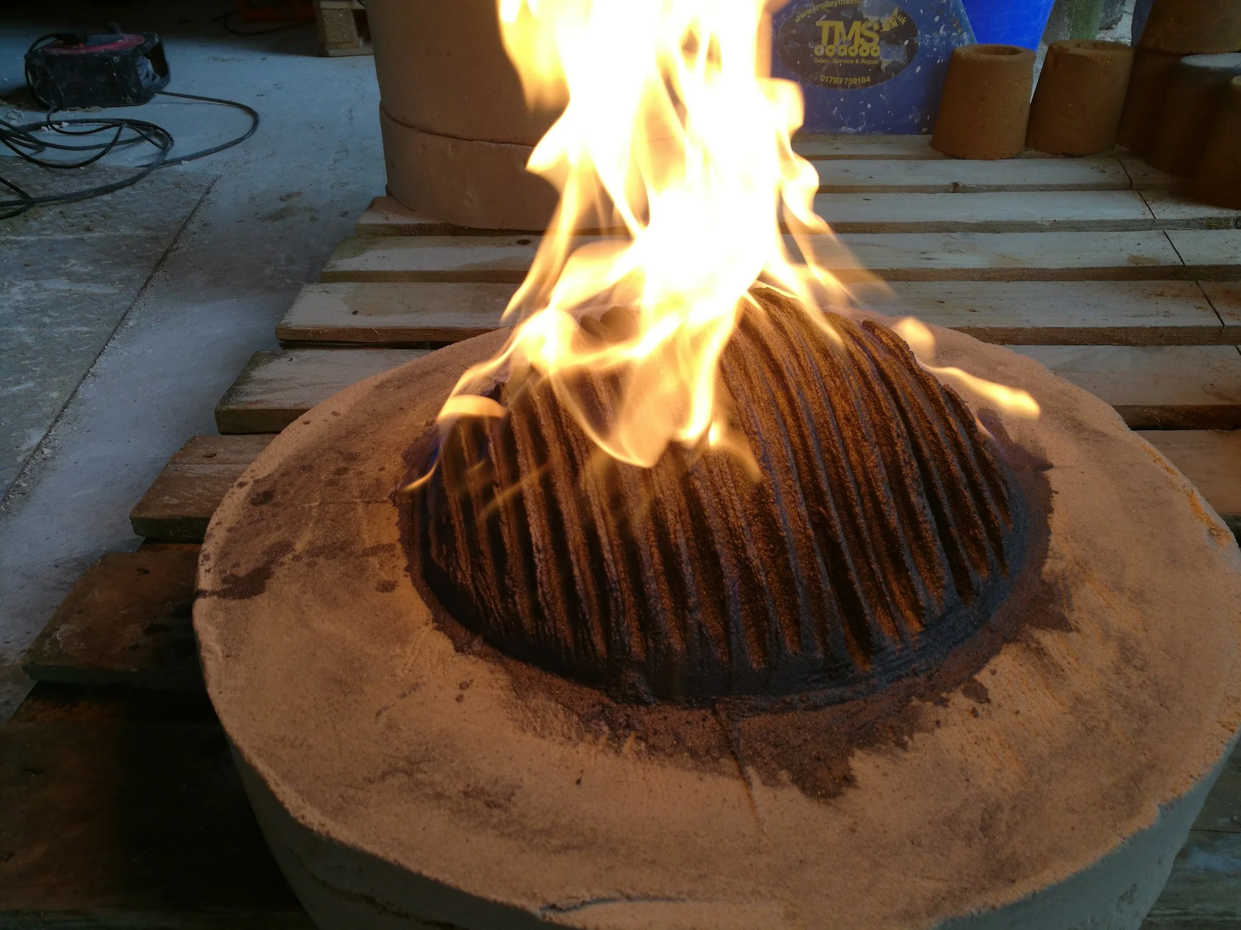 A fire burning on a stone surface in a workshop or construction area, with wooden planks and various tools and equipment in the background.