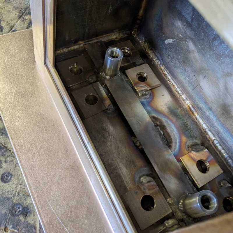Close-up of a metal fabrication with welded joints, holes, and threaded tube inserts inside a partially assembled stainless steel structure.