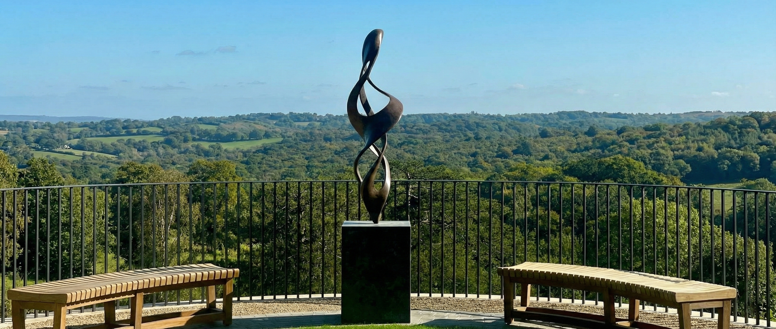 Scenic outdoor viewpoint with a modern metal sculpture on a black pedestal, overlooking lush green rolling hills, trees, and a bright blue sky with some clouds.
