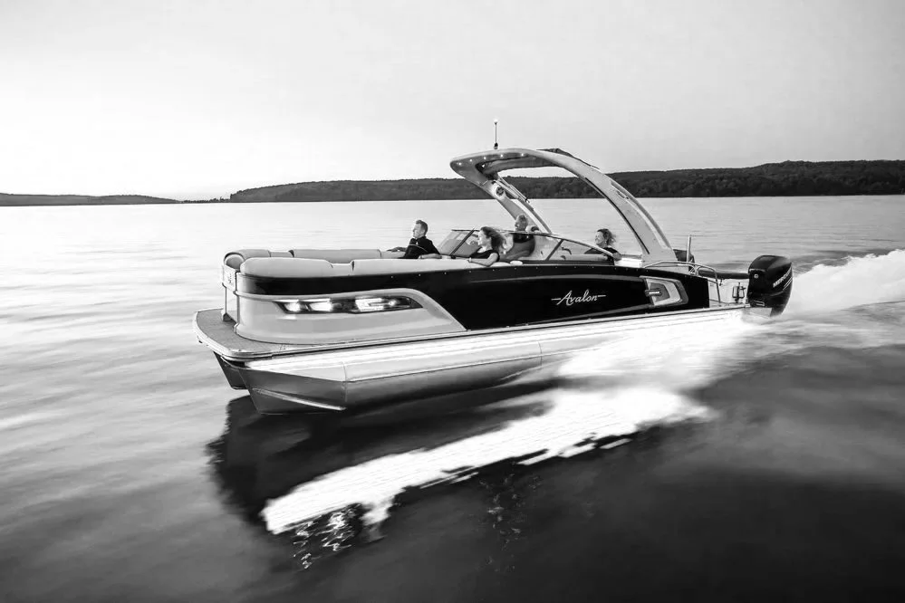 A black and white photo of a boat named Avalon speeding across a lake, with land and trees visible in the background.