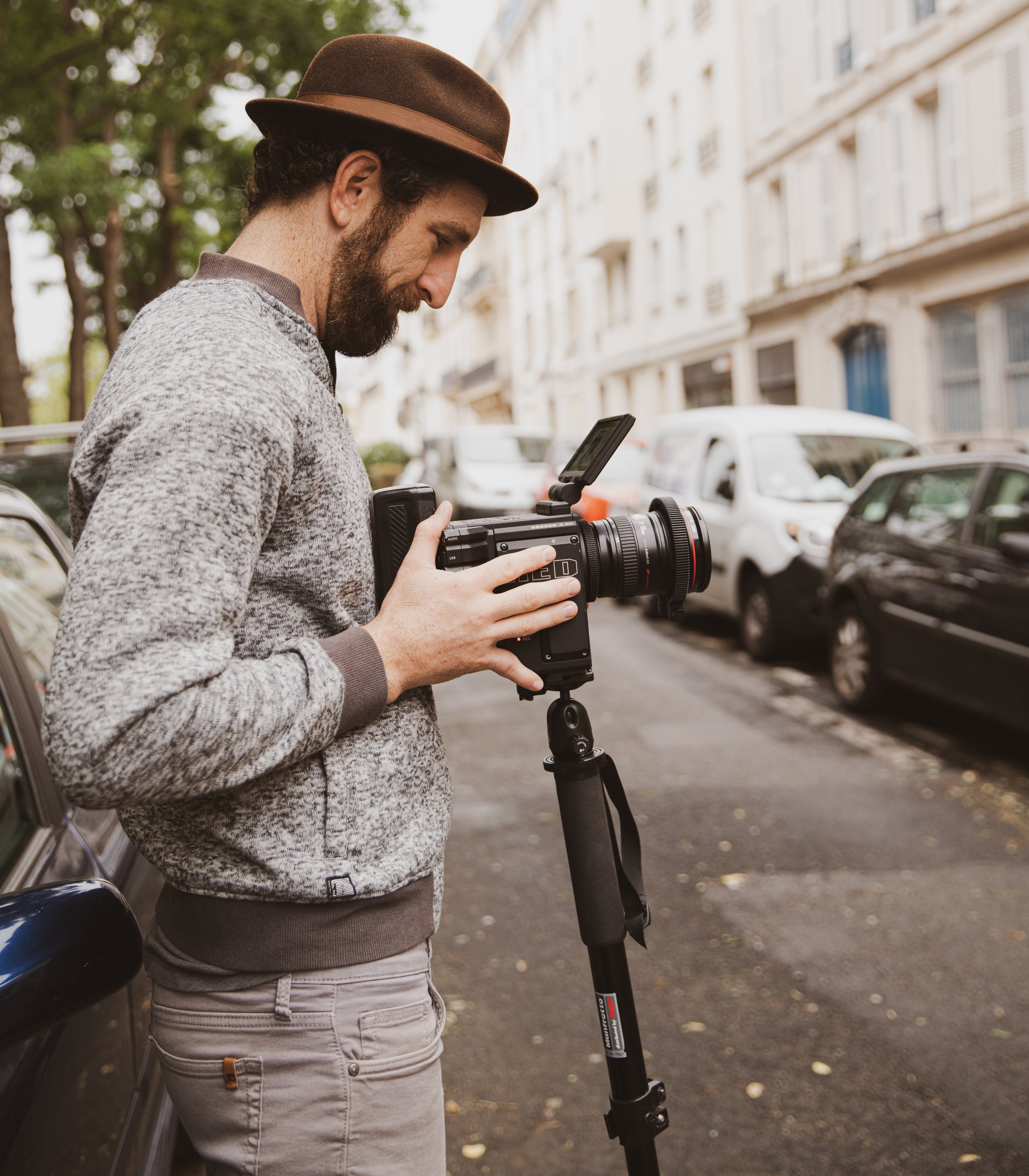 A man with a beard wearing a brown hat and a gray sweater is looking at a camera mounted on a tripod on a city street with parked cars and cream-colored buildings in the background.