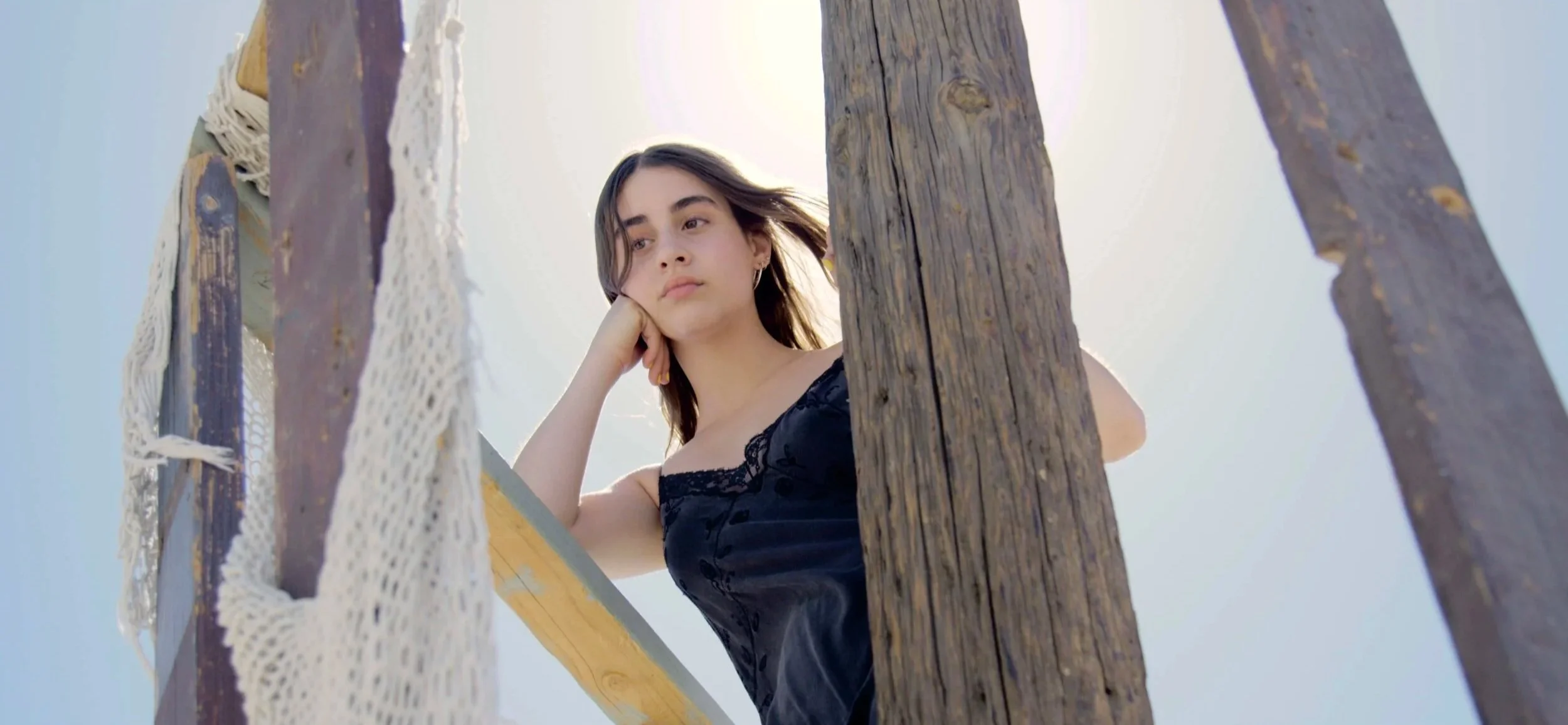 Young woman with dark hair wearing a black dress leaning on a wooden railing, looking pensively into the distance, with a clear sky in the background.