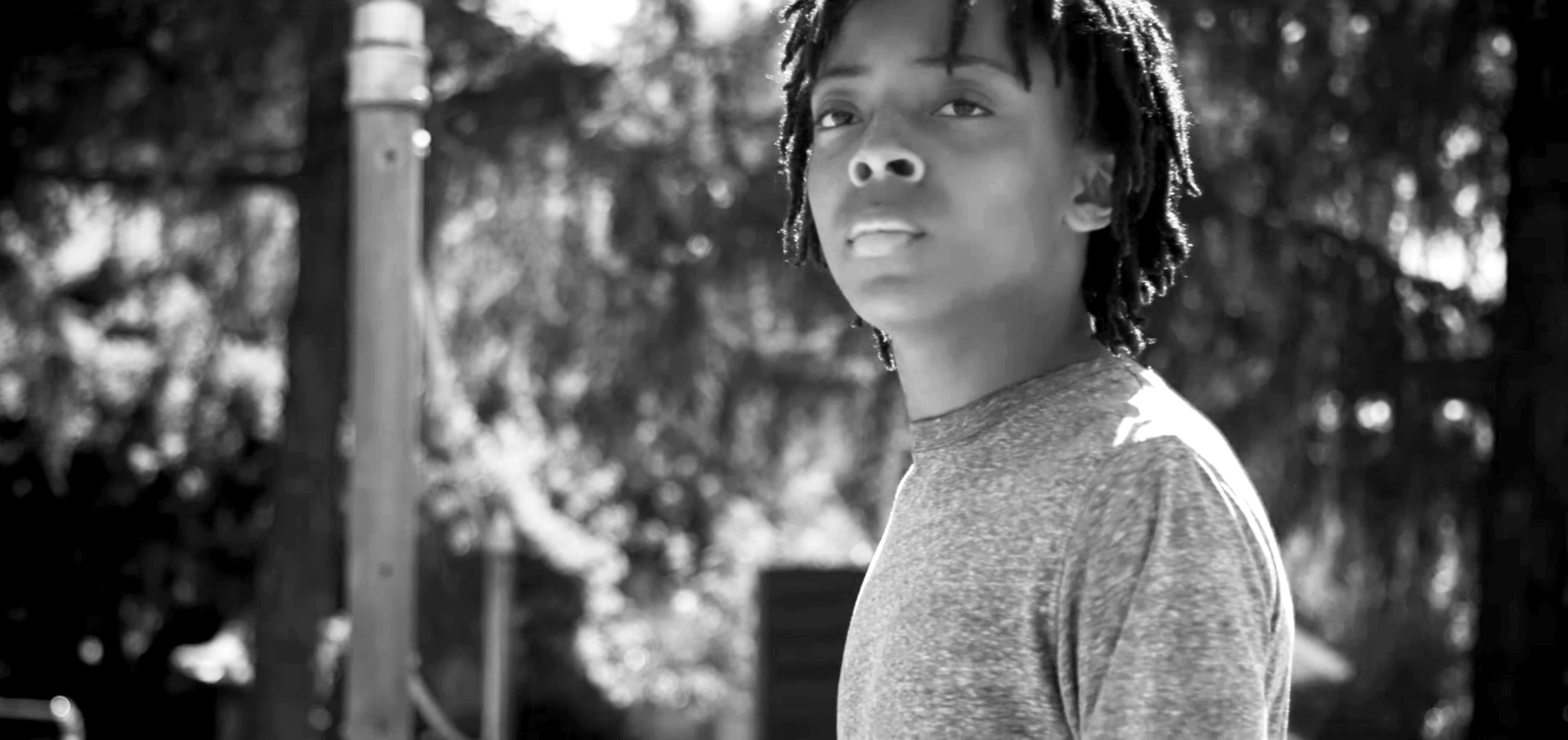 A young man with dreadlocks looking to the side outdoors, wearing a t-shirt, in black and white.