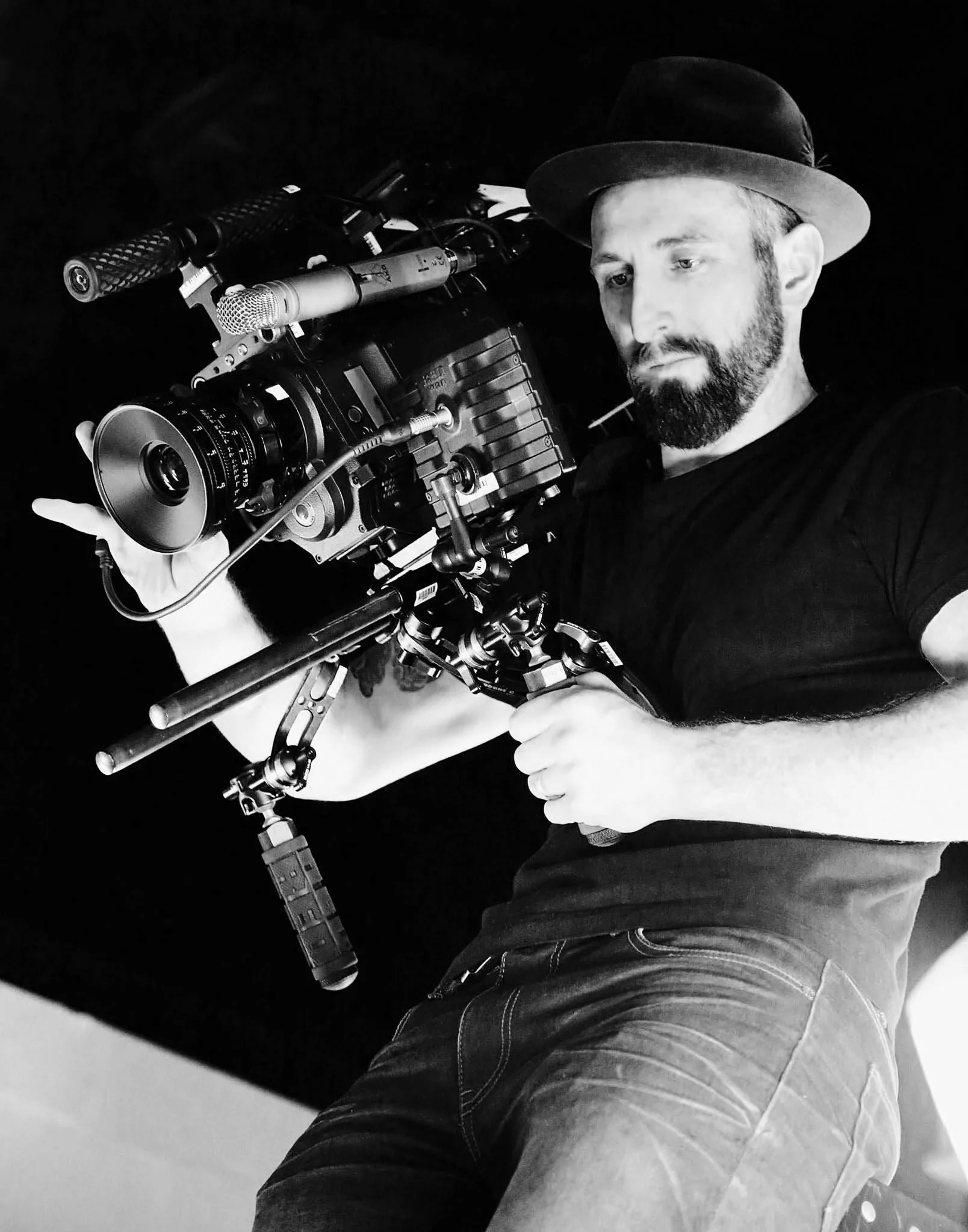 A man in a hat holding a professional film camera, aiming it as if filming, with a dark background.