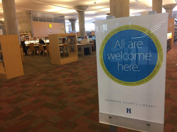 Image of a sign at the Hennepin County Library that says "All are welcome here." Via School Library Journal