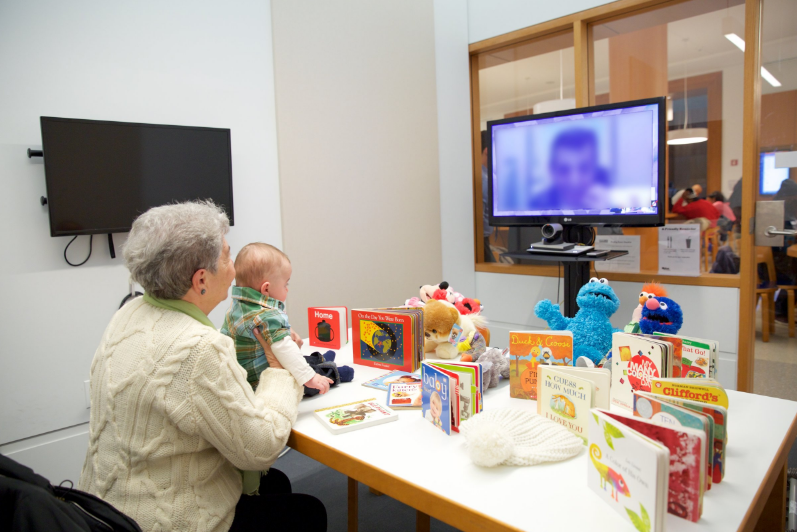 Image of an adult holding a baby up to a TV screen. The child is interacting with their incarcerated parent through the screen and there are numerous books and toys in front of the baby. Via Brooklyn Public Library