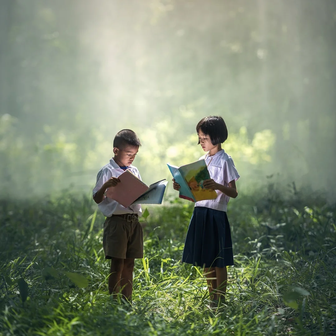 An image of two children standing in a green field while reading books.