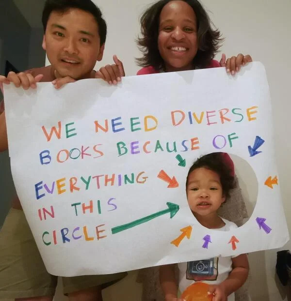 Image of young adult author Nicola Yoon and her husband holding up a sign that says, “We need diverse books because of everything in this circle.” There is a circle cut out of the sign and their daughter is standing so her face is in the circle. Photo credit: Nicola Yoon
