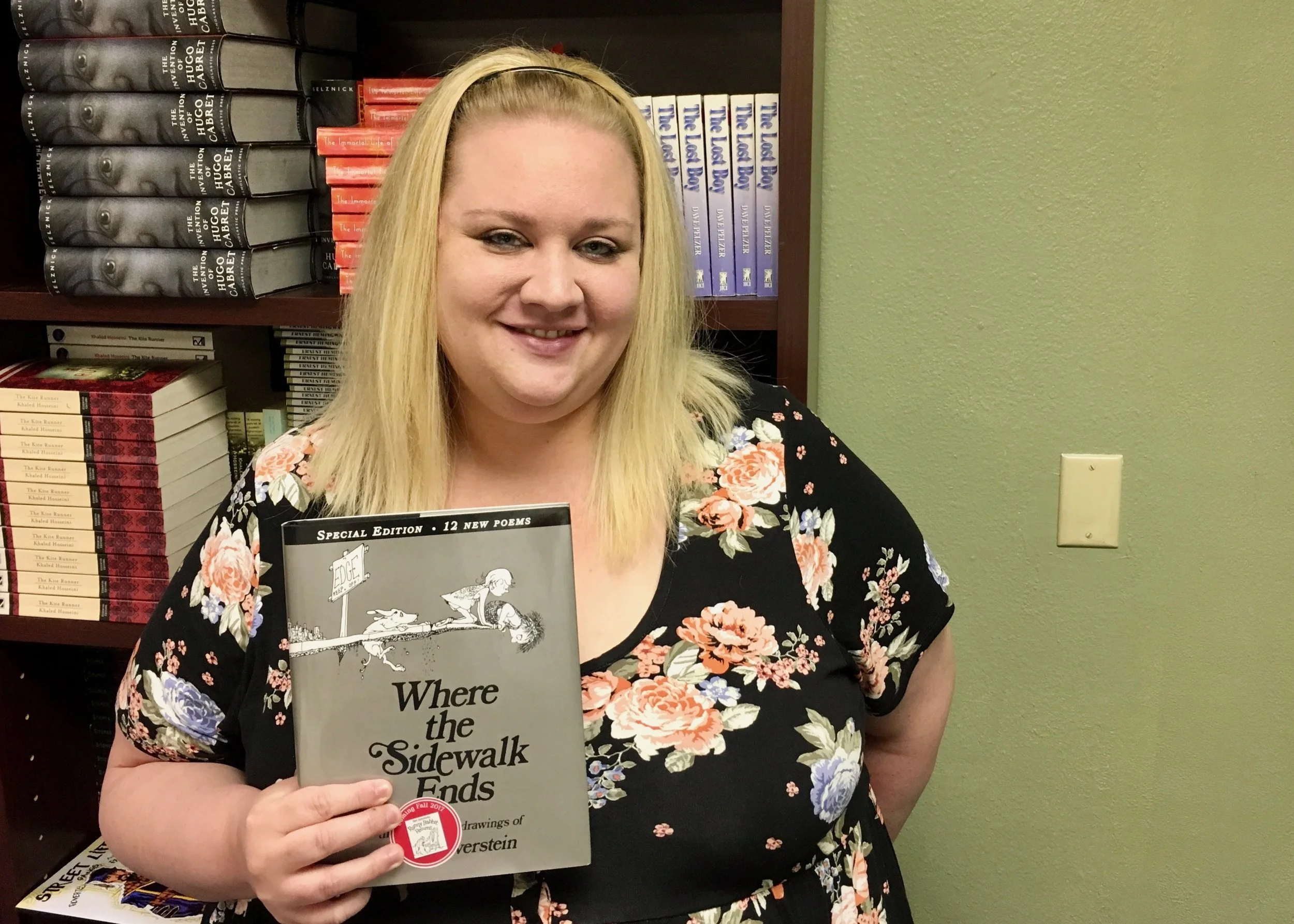 An image of Kristina Houck standing in front of bookshelves and holding up a copy of Where the Sidewalk Ends by Shel Silverstein.