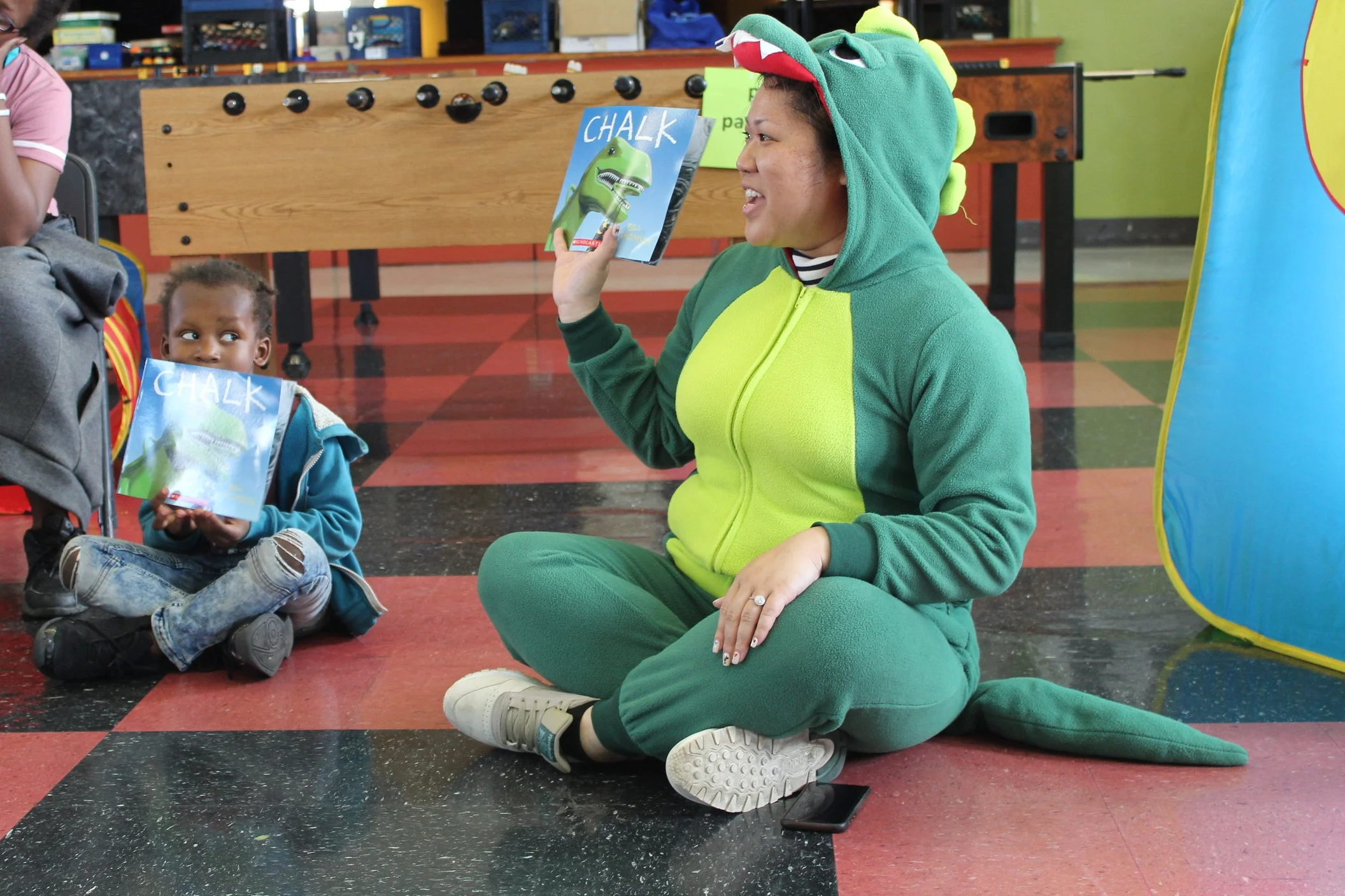 An image of Vichittra faciliting suring a Family Literacy Program session. She is wearing a dinosaur costume and holding up a book.