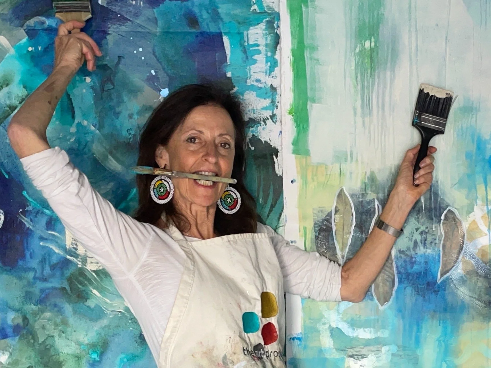 Kathy Leader in front of abstract painting with brushes in her mouth and hands