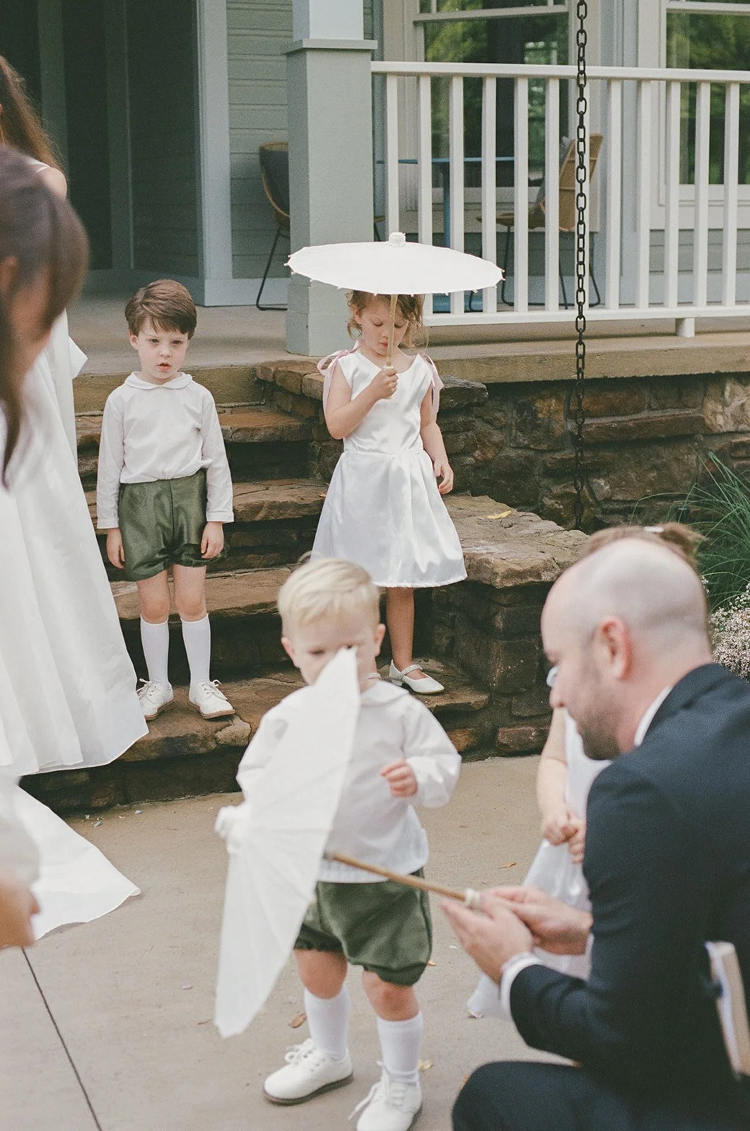 Children participating in a wedding ceremony outdoors, dressed in white and green, with some holding umbrellas, on stone steps near a porch.