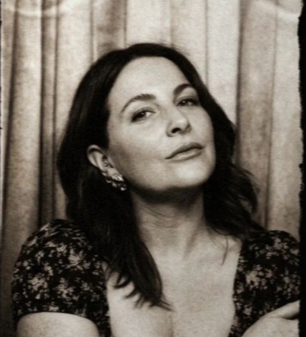 Black and white portrait of a woman with dark hair, wearing earrings and a floral dress, with a neutral expression, in front of a curtain background.