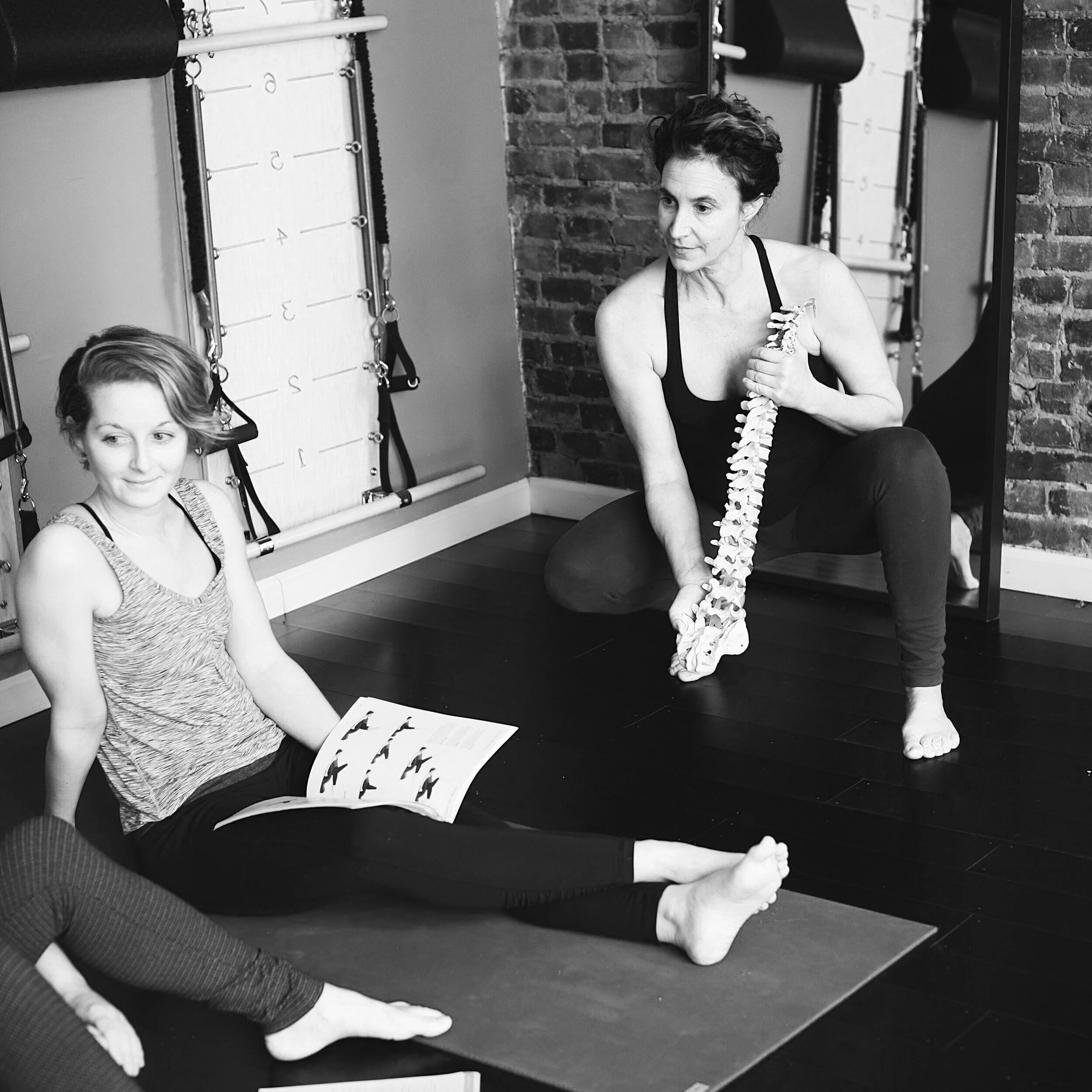 Two women in workout attire sitting on a yoga mat in a fitness studio. One is holding a spine modeland talking with the other, who is holding a booklet of exercises. The setting includes wall-mounted exercise equipment.
