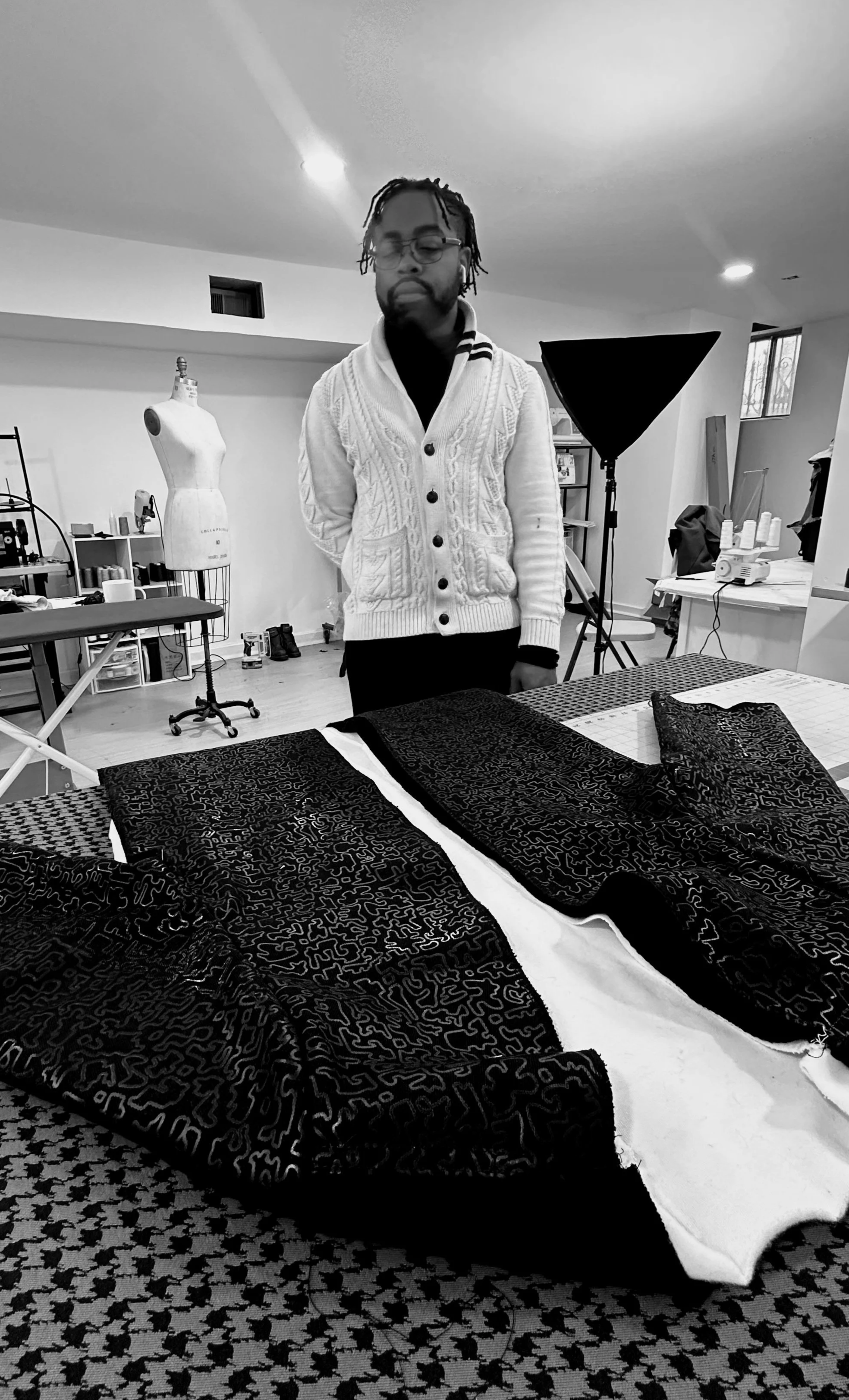 A man in glasses and a white cardigan stands behind a table with fabric swatches, in a workspace with sewing and fashion design equipment.