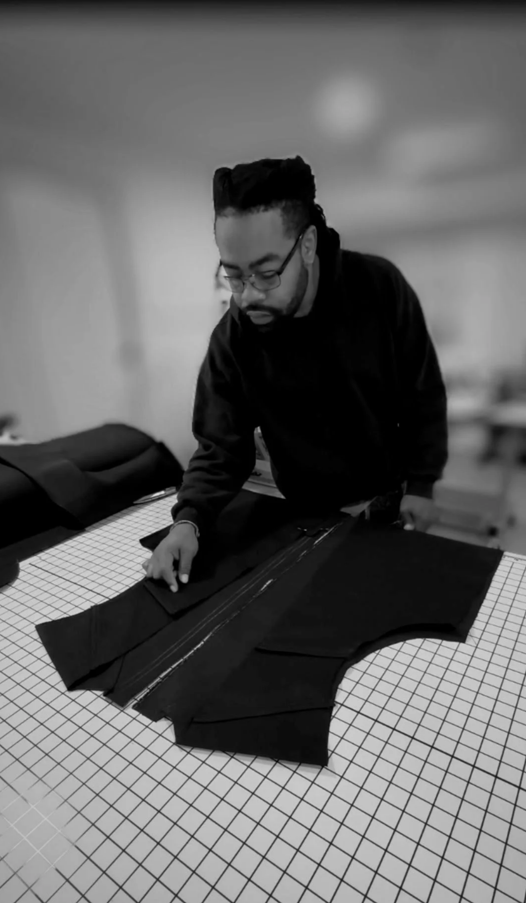 A man with glasses and dreadlocks working on black fabric laid out on a checkered cutting table.