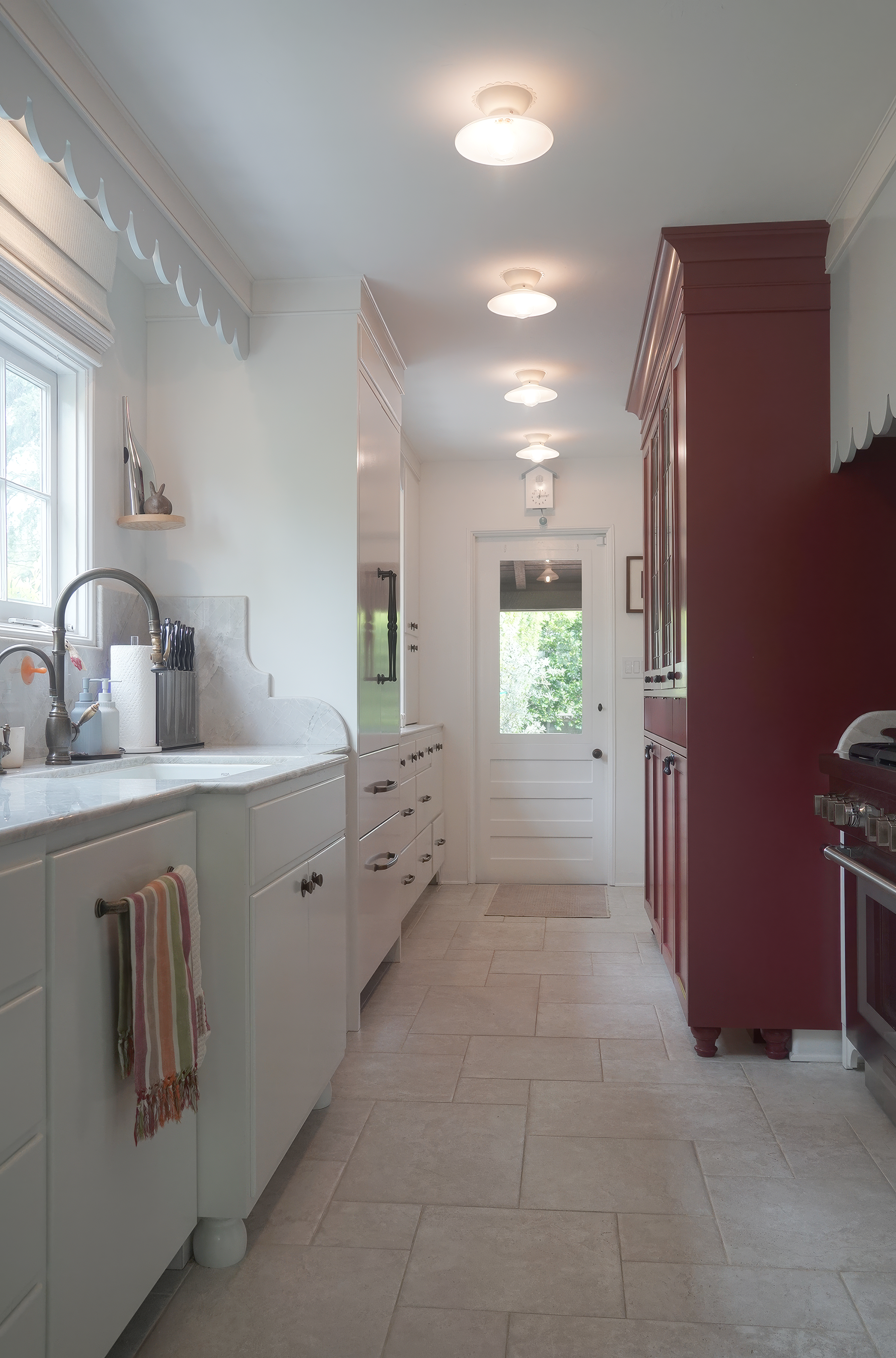 A kitchen with white cabinets, a marble countertop, a stainless steel refrigerator, a window with a valance, and a red pantry cabinet. There are ceiling lights and a door leading outside, with trees visible through the window.