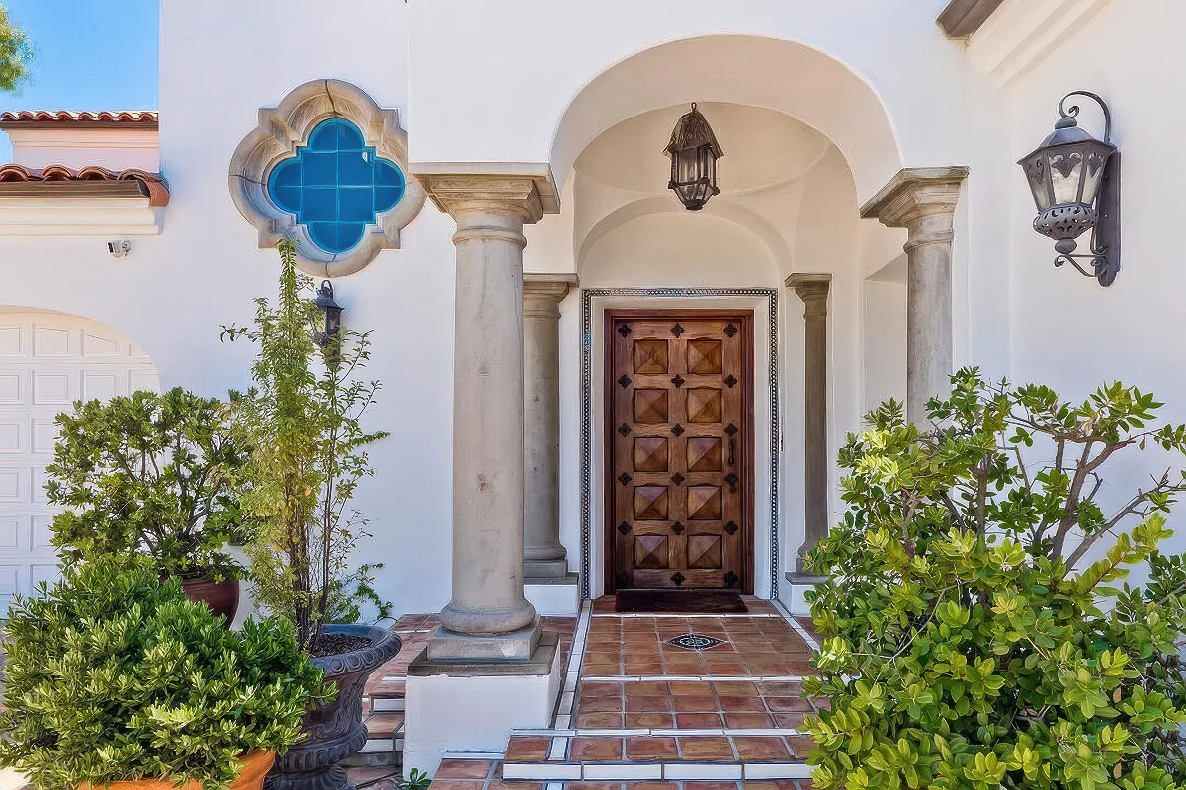 Front entrance of a house with a wooden door, stone columns, black lanterns, lush green plants, and a tiled walkway.