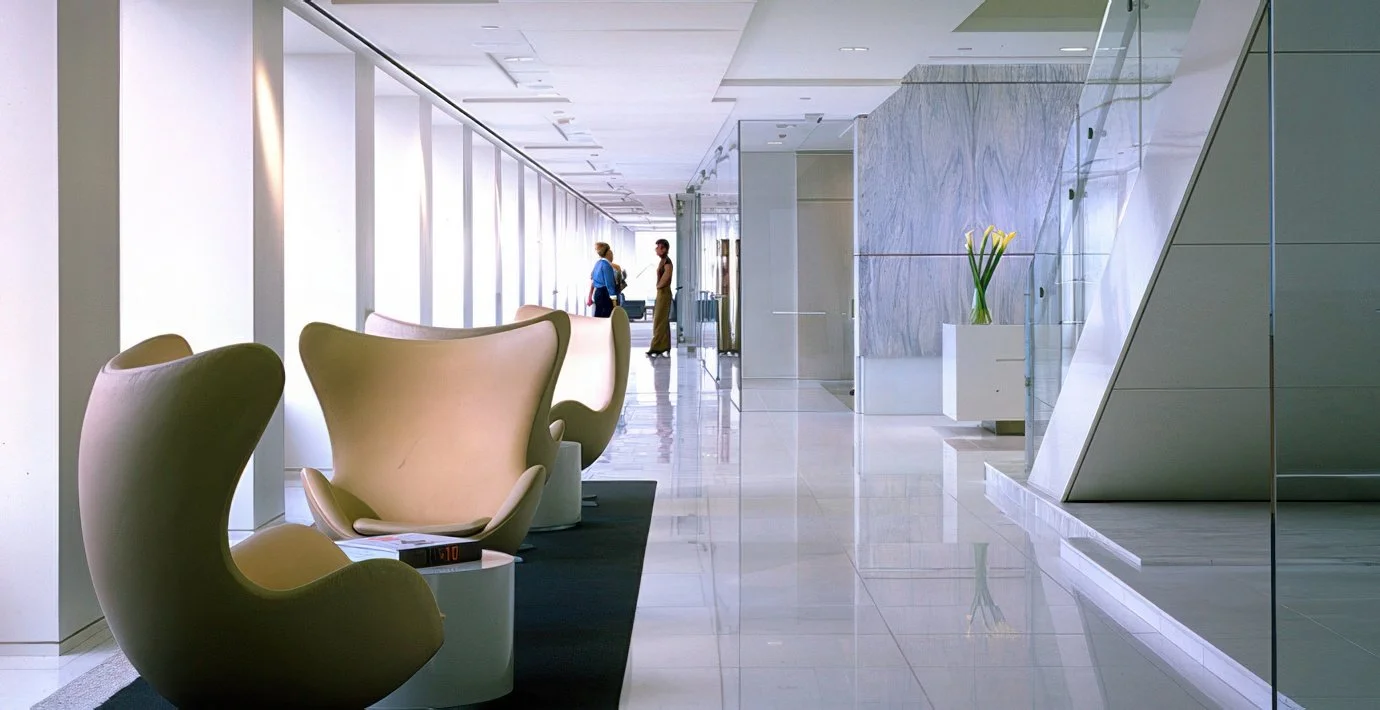 Modern hotel lobby with beige Egg chairs, a black rug, a large mirror wall, and a Vanda orchid in a white pot, with two people talking in the background.