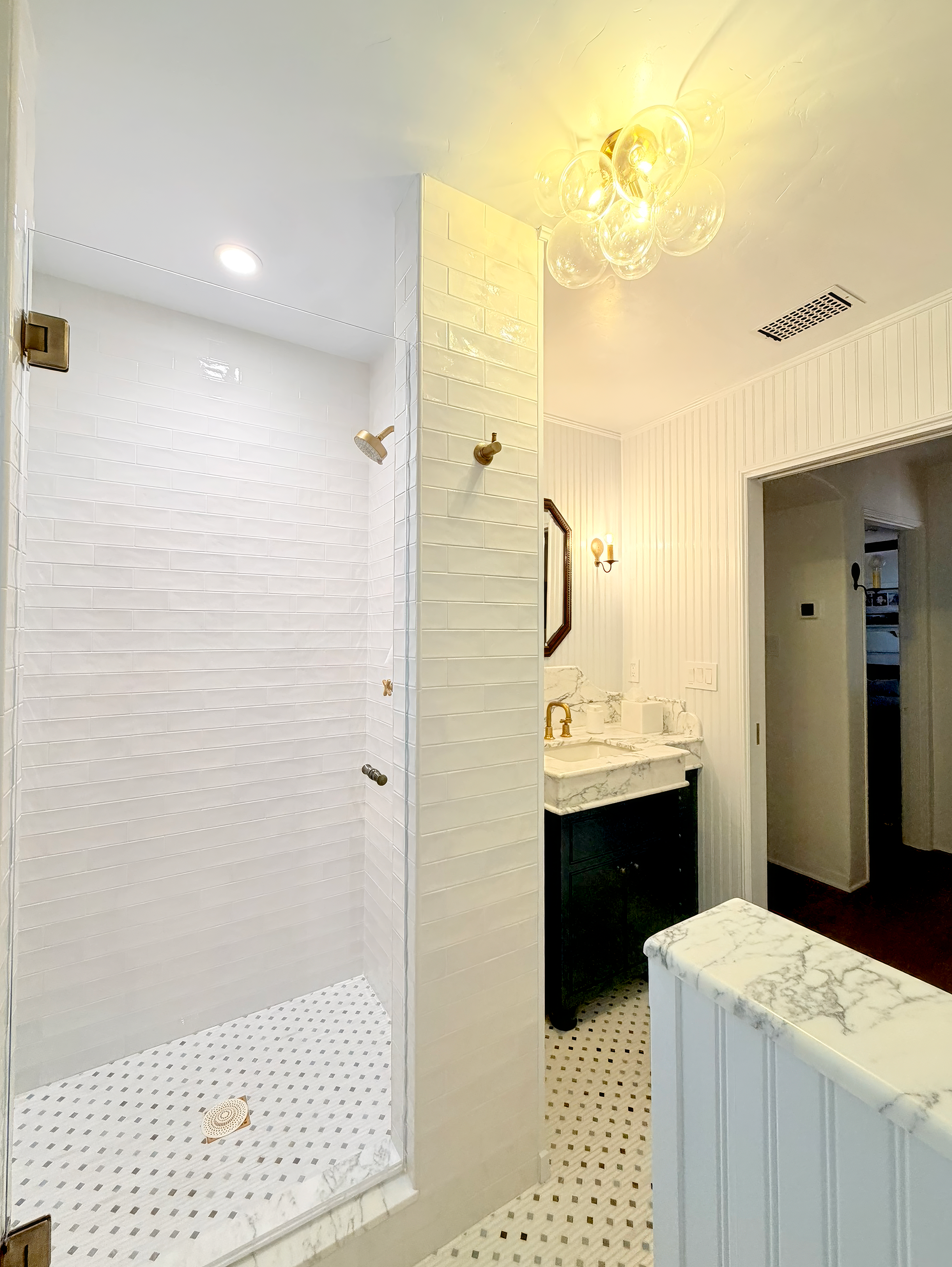 A bathroom with a walk-in shower on the left, featuring white subway tile walls and a marble threshold, and a vanity on the right with a marble countertop, black cabinet, and gold fixtures, illuminated by a modern chandelier and wall sconce.