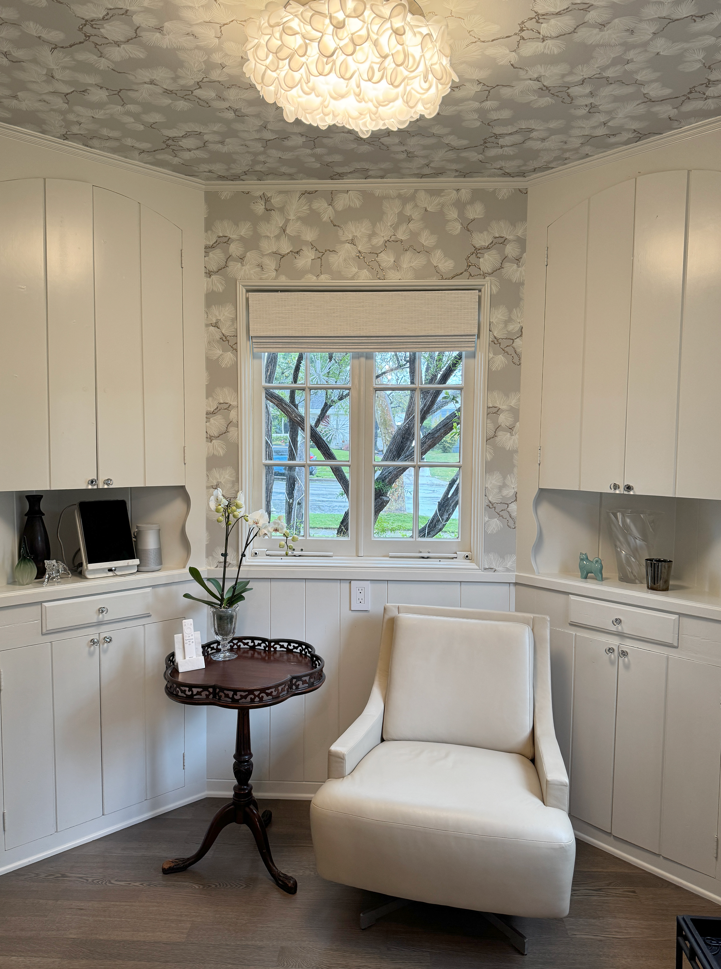A cozy corner of a room with white built-in cabinets, a beige armchair, and a small wooden table with a decorative tray. A large window with a beige Roman shade shows trees and a street outside. The ceiling has floral patterned wallpaper and a white 