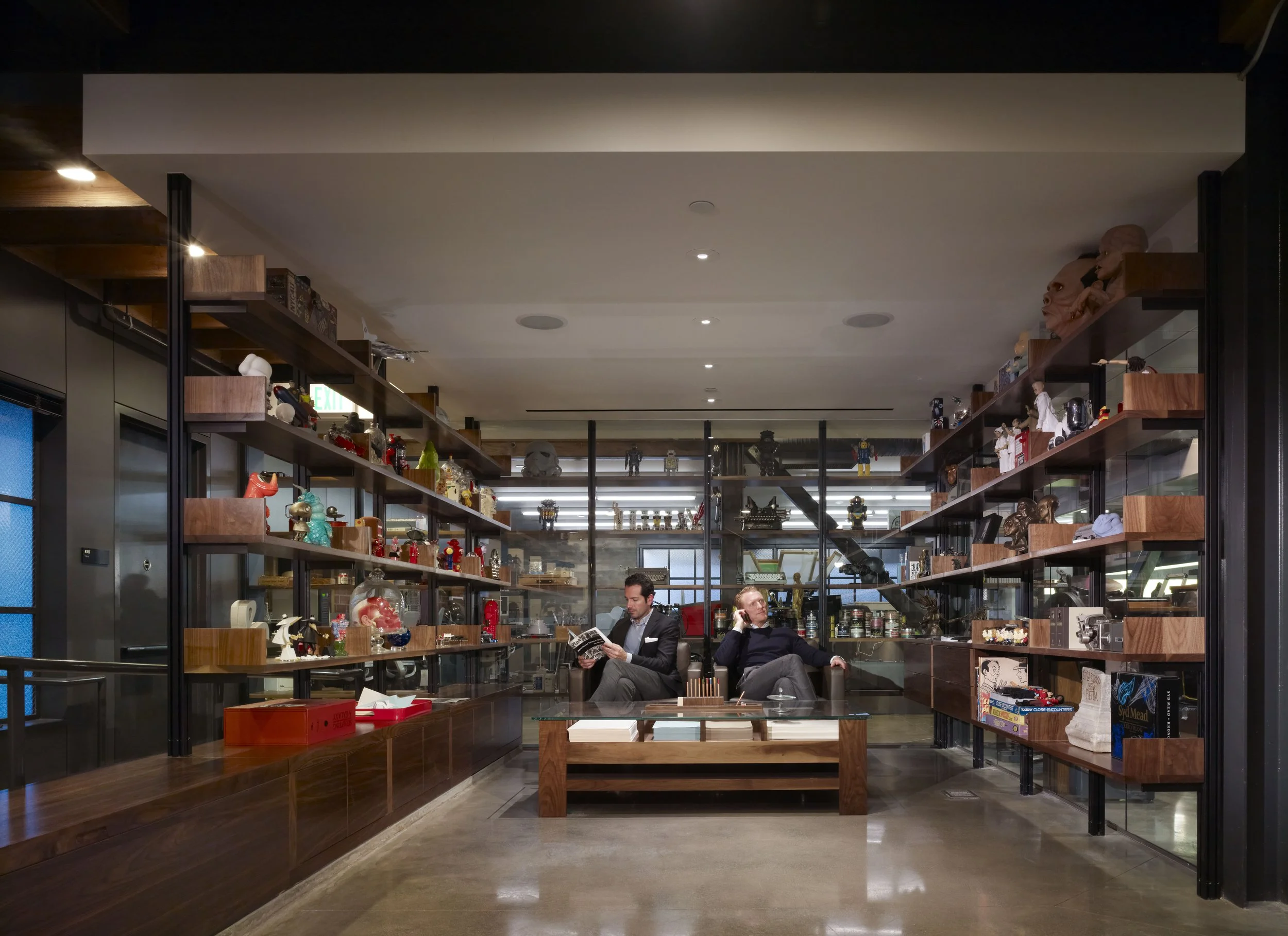 A bookstore or shop with shelves of books and collectibles, two men sitting and talking, one reading a book, inside a well-lit space with dark wood furniture and decorative items.