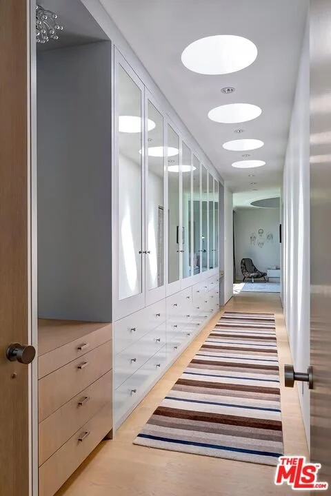 Bright hallway with white built-in cabinets with glass doors, a striped rug on light wooden floor, circular ceiling lights, and a sitting area at the end with a black chair and wall art.