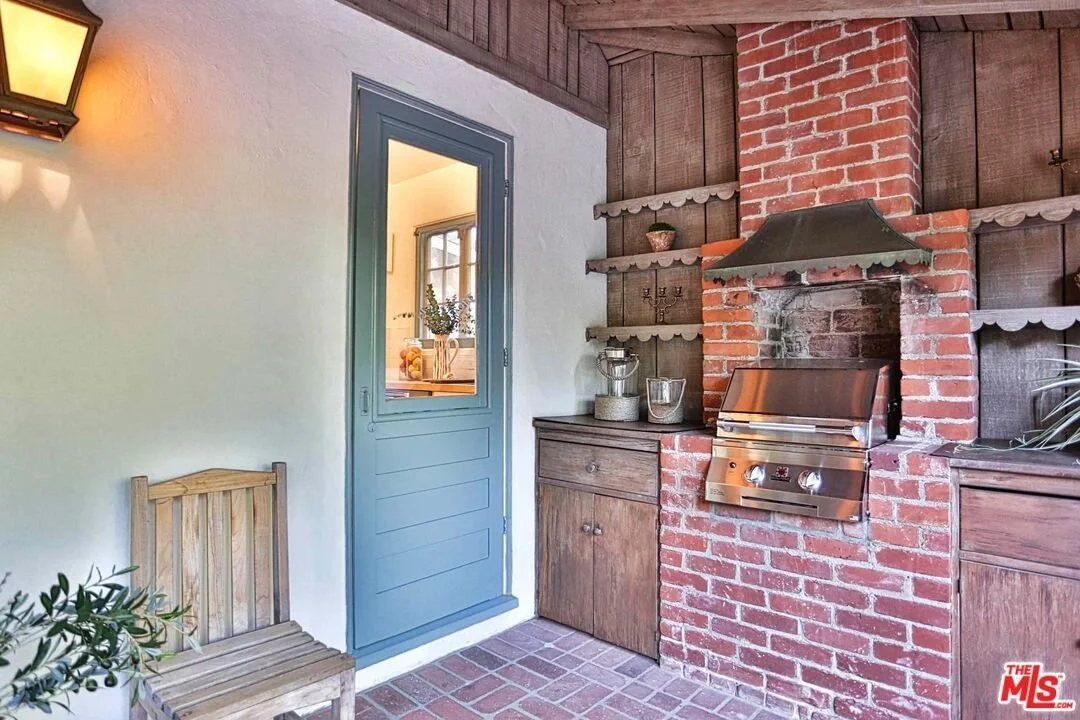 An outdoor brick fireplace with a stainless steel grill, open shelves with decor, a small wooden cabinet, and a blue door leading inside, with a wooden chair and a potted plant nearby.