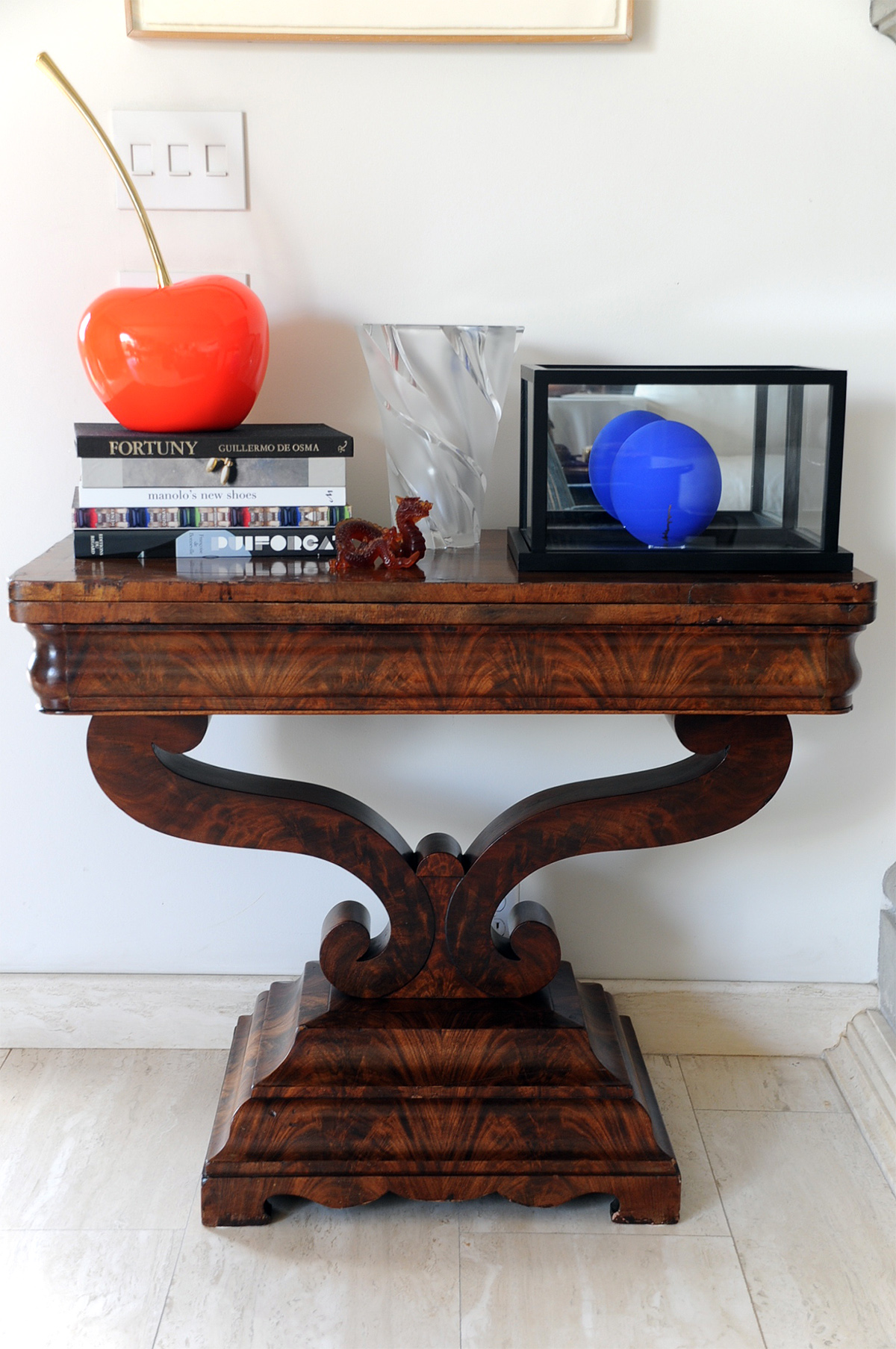 Wooden table with a stack of books, a glass vase, and decorative objects on top, set against a white wall.