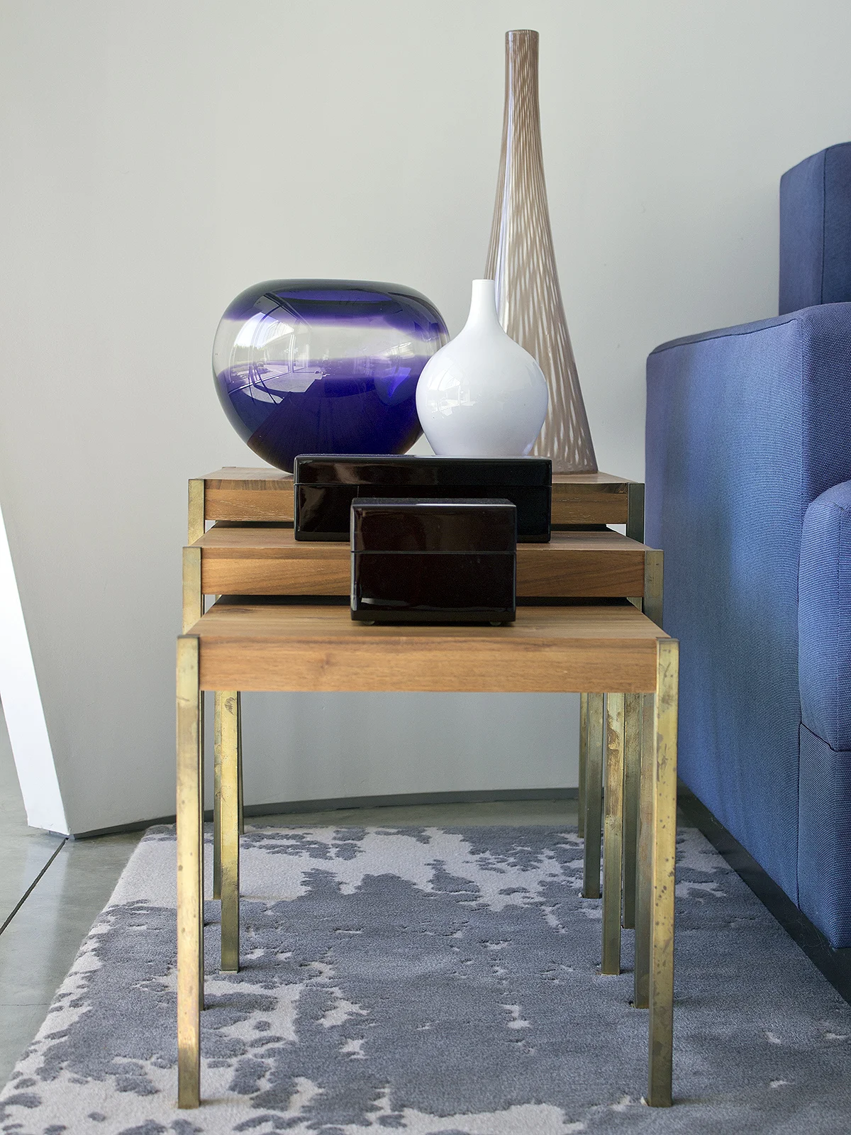 A wooden side table with brass legs holding decorative vases, including a purple and clear glass vase, a white ceramic vase, and a tall, thin wooden lamp, next to a blue upholstered sofa.