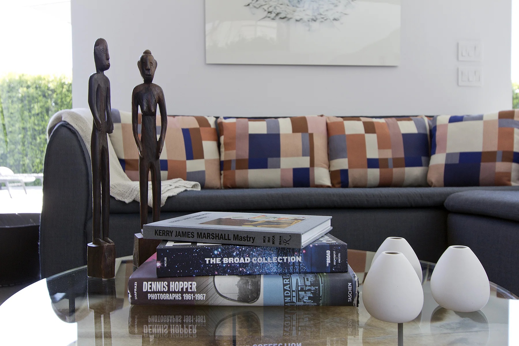 Living room with a stack of art books on a glass table, decorative white vases, two wooden figurines, a dark gray sofa with a colorful geometric patterned cushion, and abstract artwork on the white wall behind.