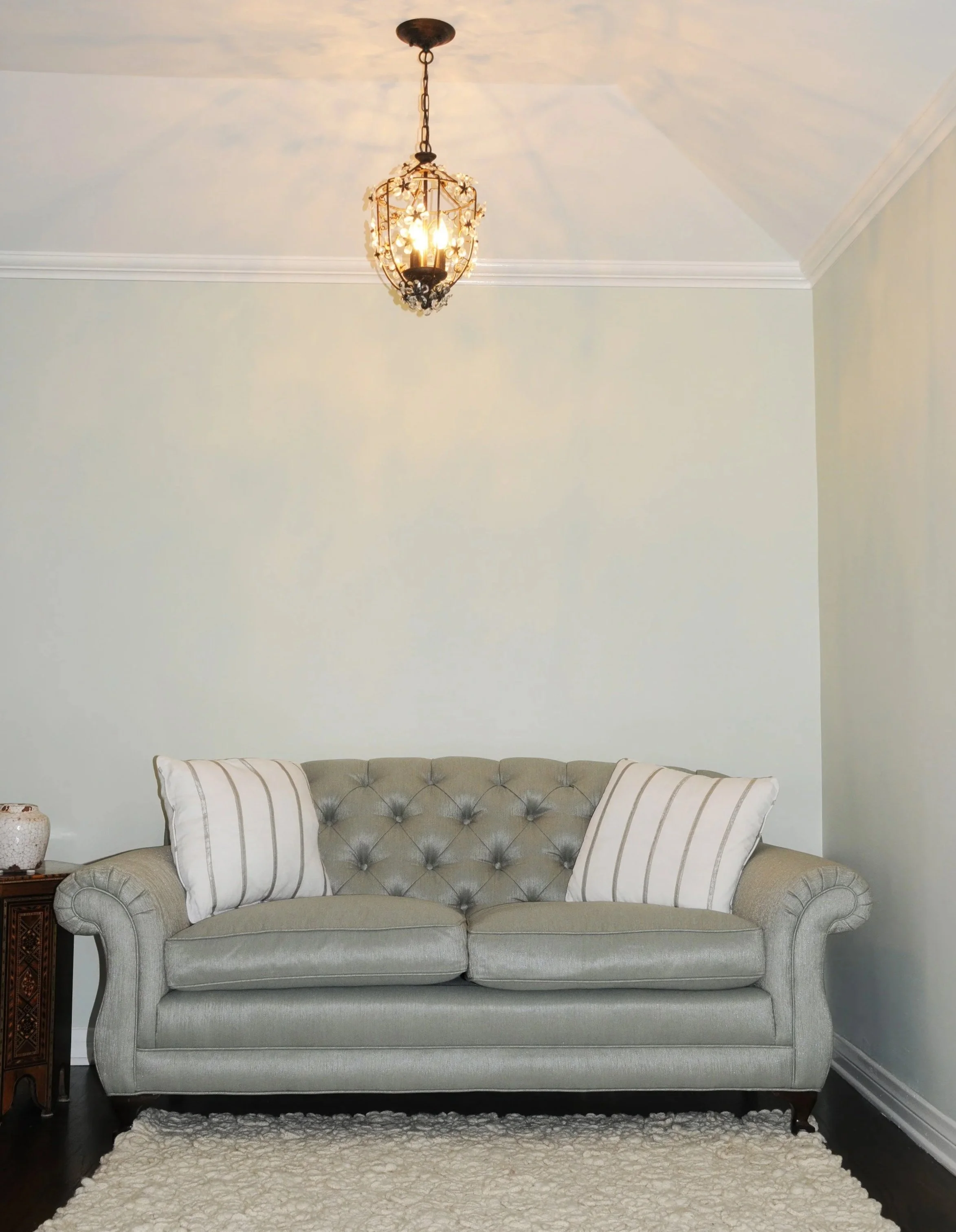 Living room with a gray tufted sofa, two white striped pillows, a cream-colored shaggy rug, a wooden side table, a ceramic vase, a chandelier, and white walls with crown molding.