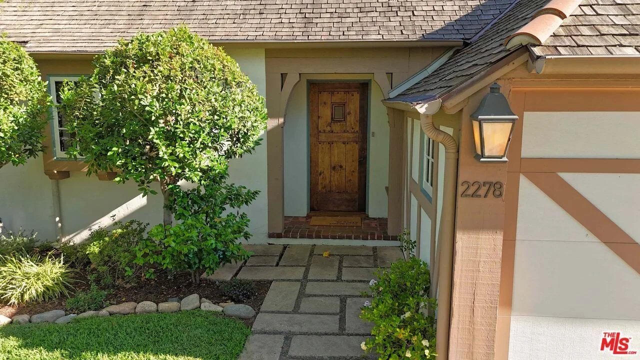 Front entrance of a house with a wooden door, stone pathway, manicured lawn, green shrub, and a wall-mounted lantern light.