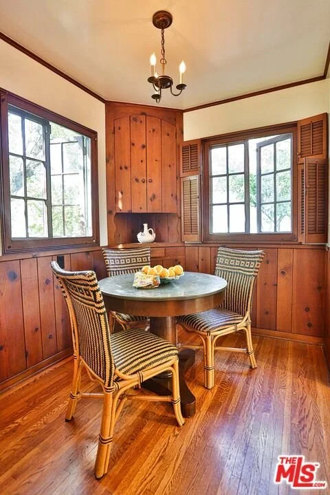A rustic dining nook with two large windows, wood-paneled walls, a round table, three wicker chairs, and a chandelier with three candle-shaped lights.