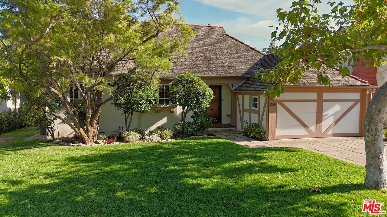 A house with a brown shingle roof, white walls, and a two-car garage with wooden trim. There is a sidewalk leading to the front door, surrounded by green grass and trees with foliage.