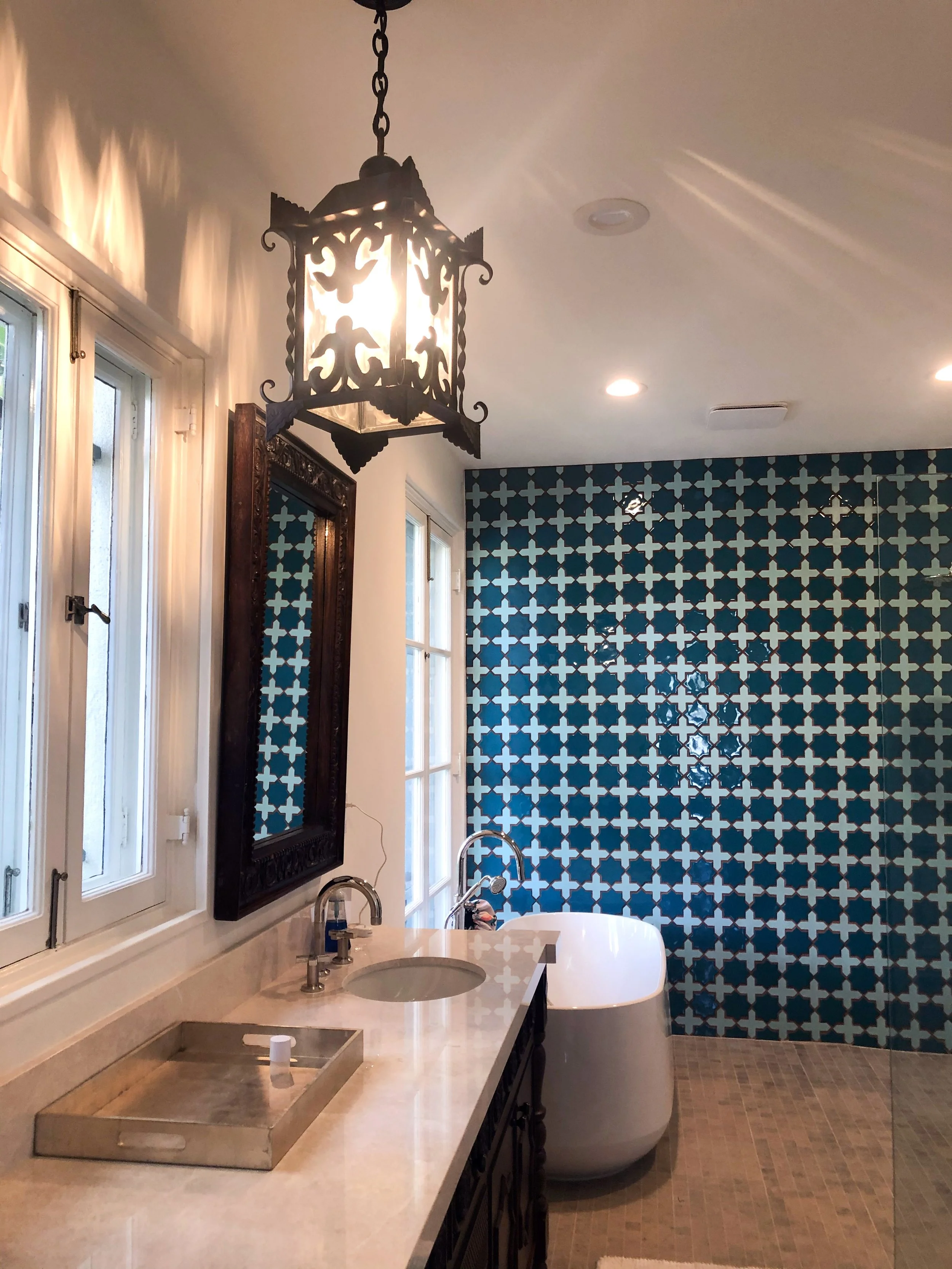 Bathroom interior with blue geometric patterned tiled wall, white bathtub, vanity with marble countertop, mirror, and light fixture, double-hung window, and wood-floored area.