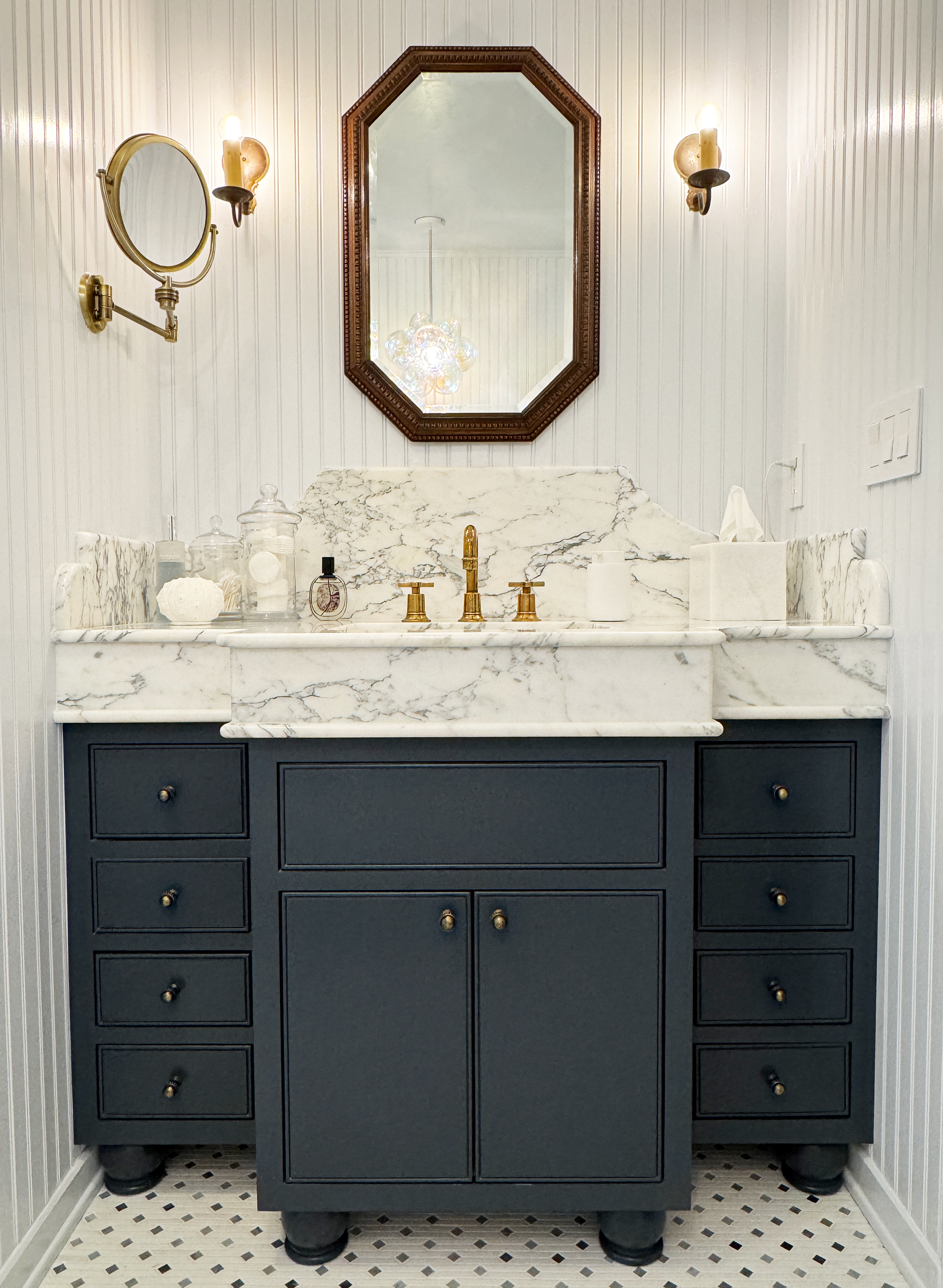 A bathroom vanity with a marble countertop and a large mirror, featuring black drawers with brass knobs, a brass faucet, and decorative items including jars, a small bottle, a bowl, a tissue box, and a soap dispenser. The wall has white beadboard pan
