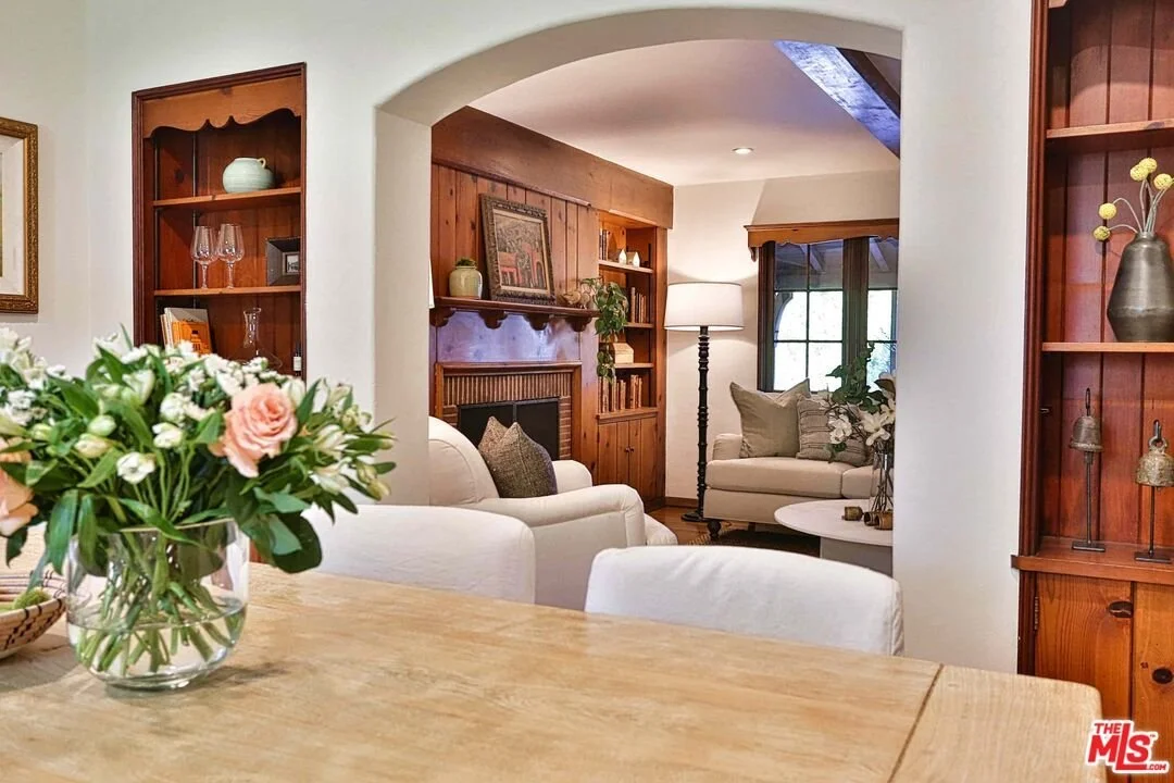 Living room with white sofas, wooden built-in shelves, fireplace, and large windows, viewed from a wooden dining table with a vase of flowers.