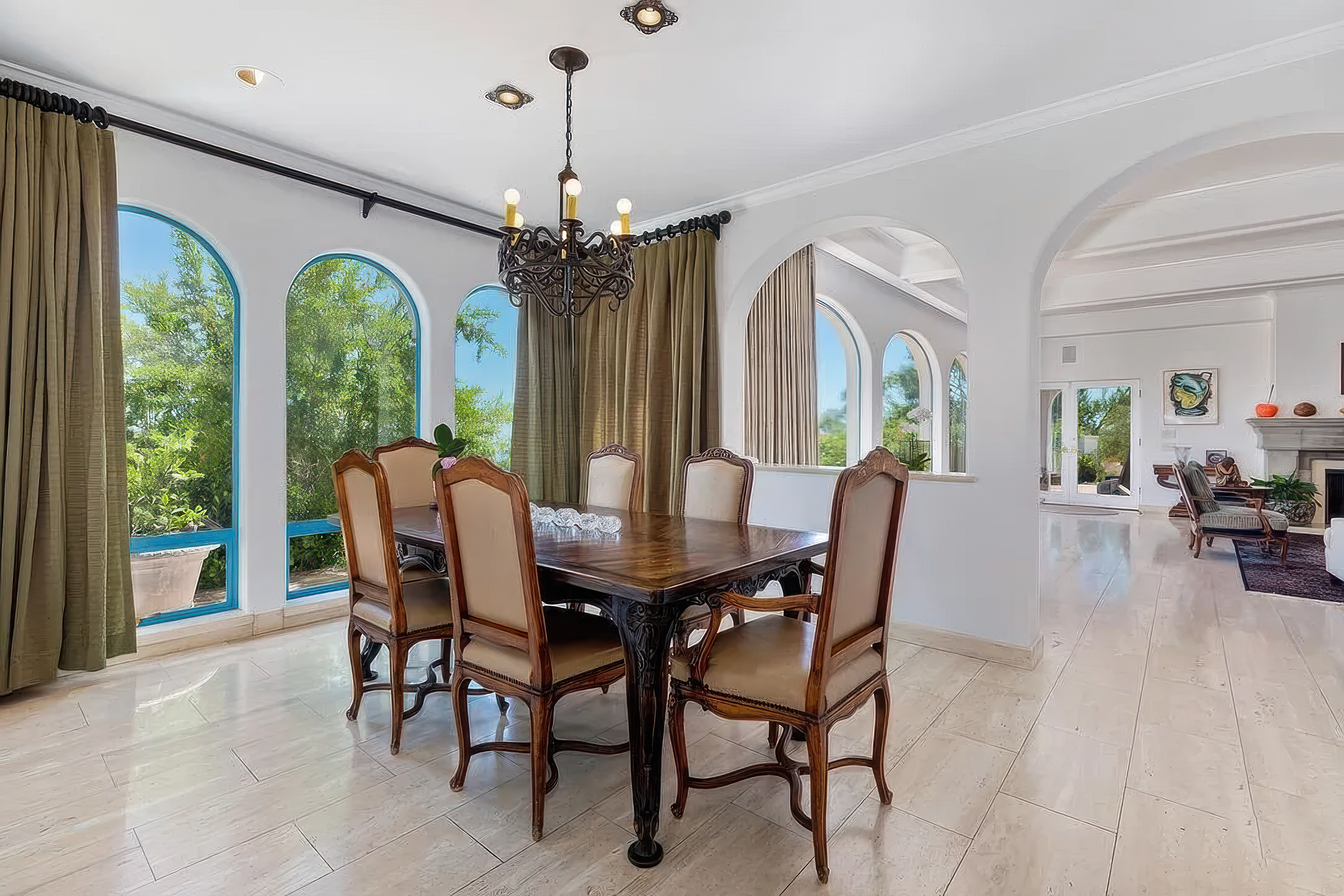 A dining room with a large rectangular wooden table surrounded by eight upholstered chairs. The room has large arched windows with beige curtains, allowing ample natural light. A black ornate chandelier hangs from the white ceiling. Visible outside t