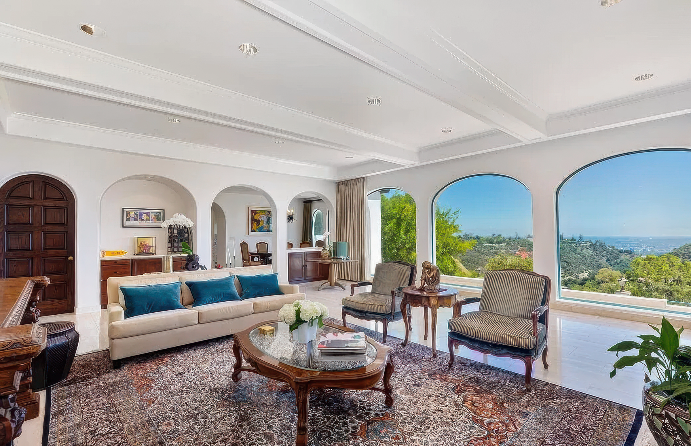 Living room with white walls, large arched windows showing greenery and a view of the ocean, various furniture including a sofa with blue pillows, armchairs, and wooden tables, decorative art on the walls, a patterned rug, and a ceiling with recessed