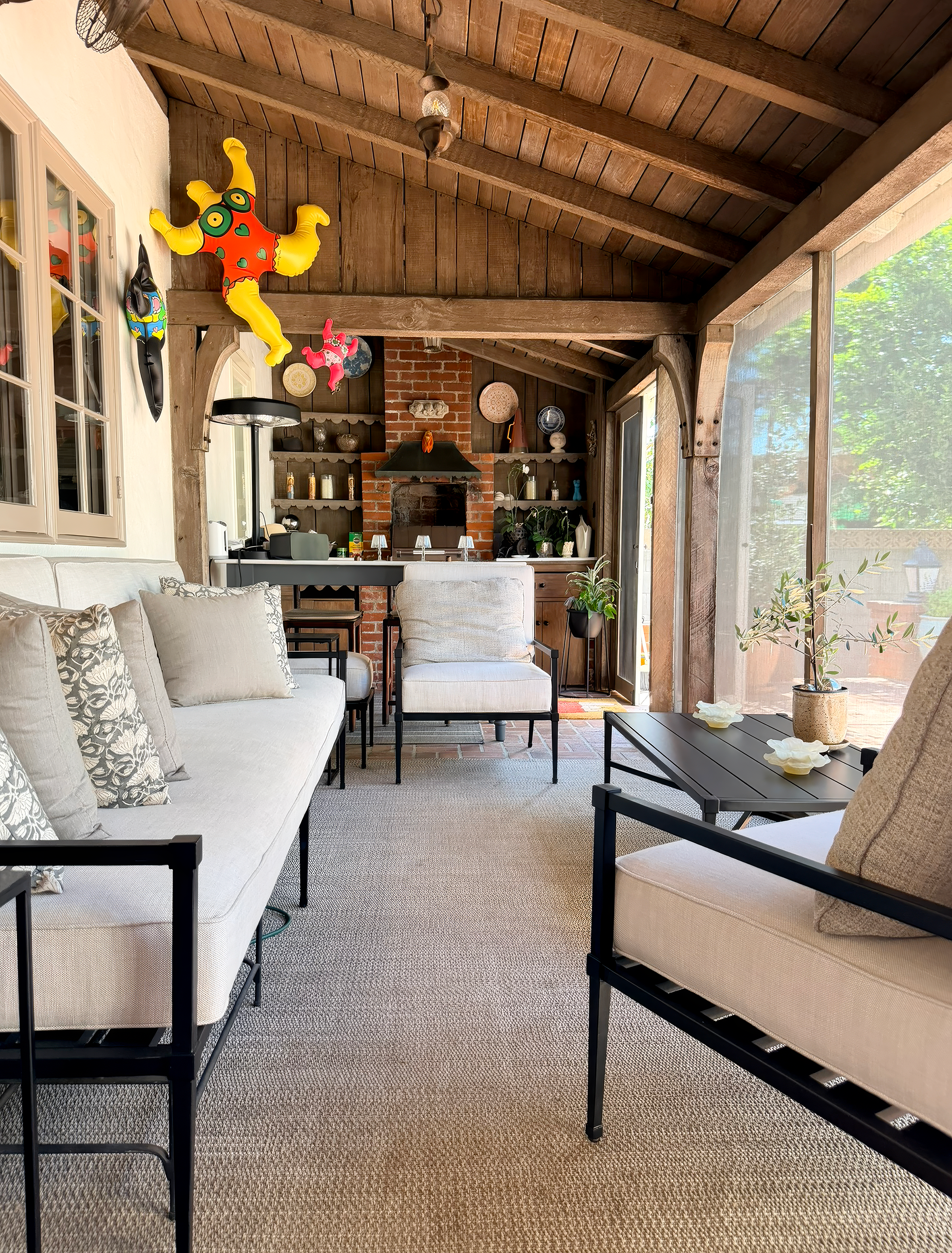 A cozy indoor seating area with white sofas, beige armchairs, and black metal-framed chairs arranged around a coffee table with white decorative objects. The space features wooden beams on the ceiling, a brick fireplace, and shelves with various deco