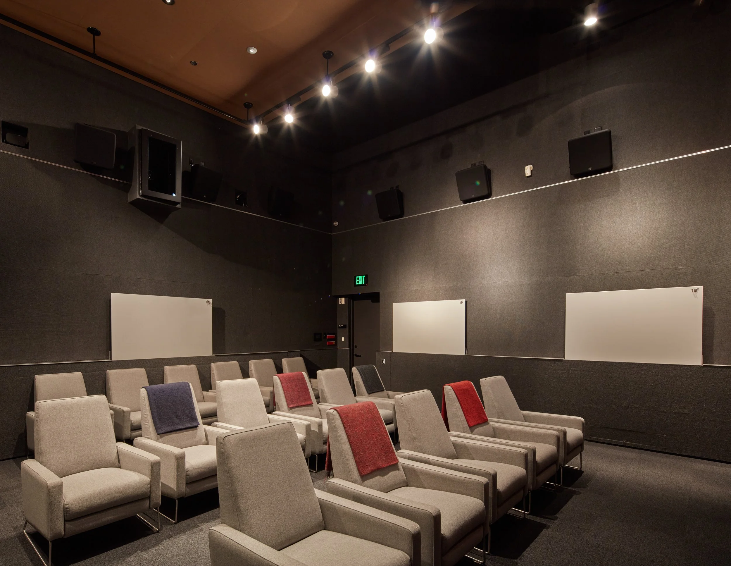 Empty auditorium with beige chairs and red towels, black walls, multiple speakers, a lighting track with several lights on the dark ceiling, whiteboards on the walls, and an illuminated green exit sign.