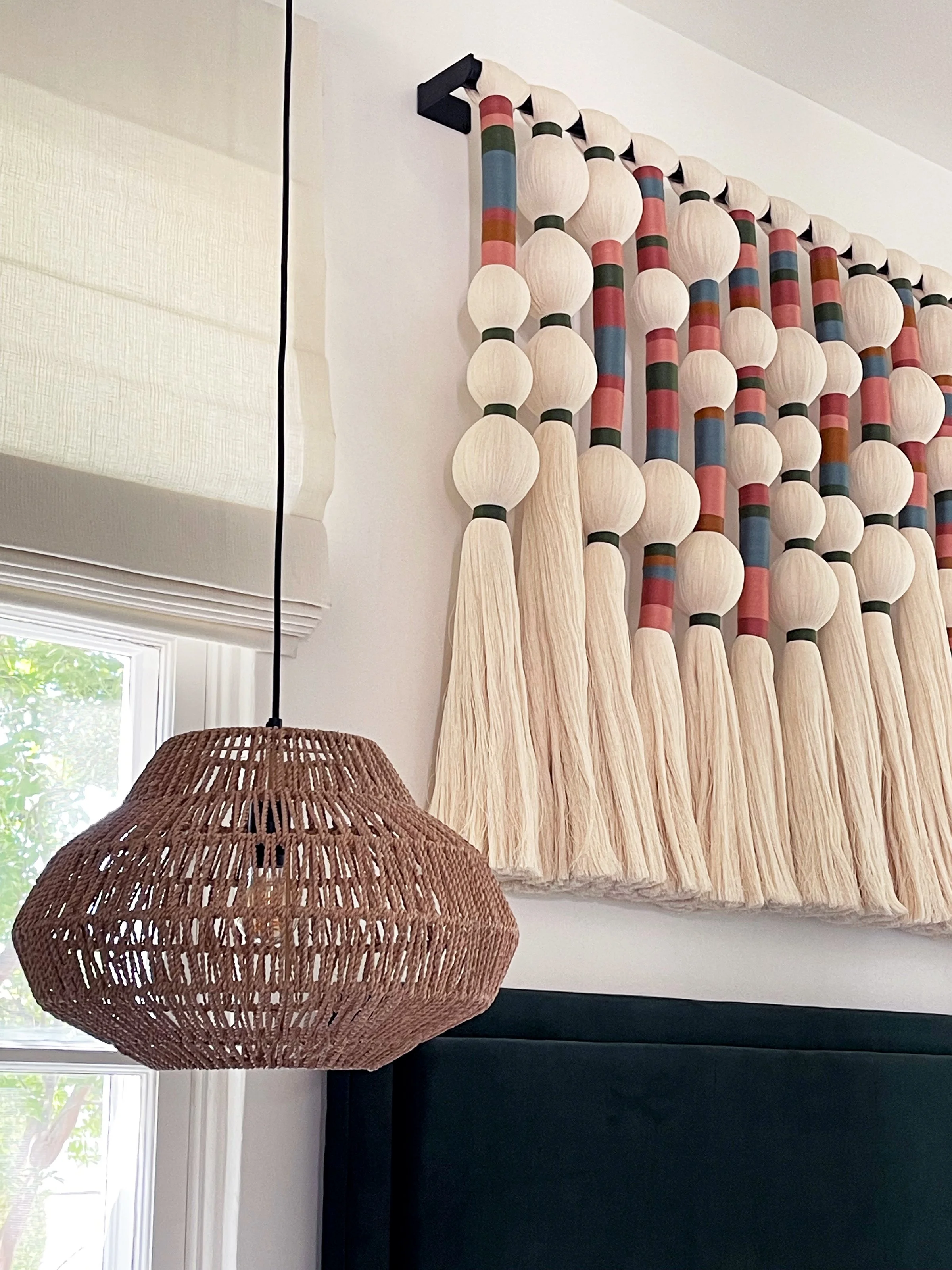 Indoor decor featuring a hanging wicker lamp and a fabric curtain with wooden beads and colorful painted sections, near a window with white trim.