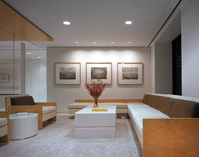 Modern waiting area with minimalist furniture, including a long beige sofa, a white coffee table with a vase of dried flowers, and framed black-and-white artwork on the wall.