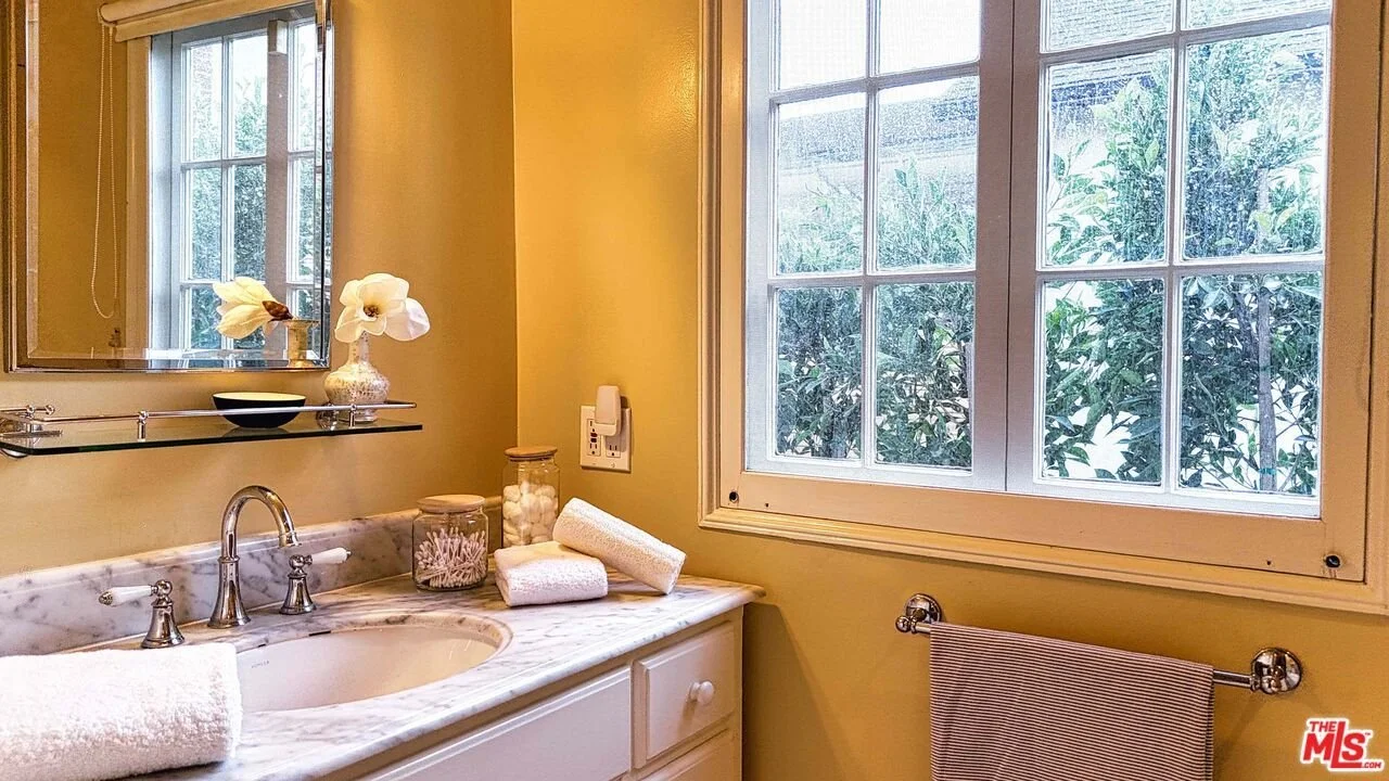 A bathroom vanity area with a marble countertop, a sink with silver fixtures, and a large window showing greenery outside. The wall above the sink has a mirror and a glass shelf holding a small black bowl and a vase with white flowers. There are towe