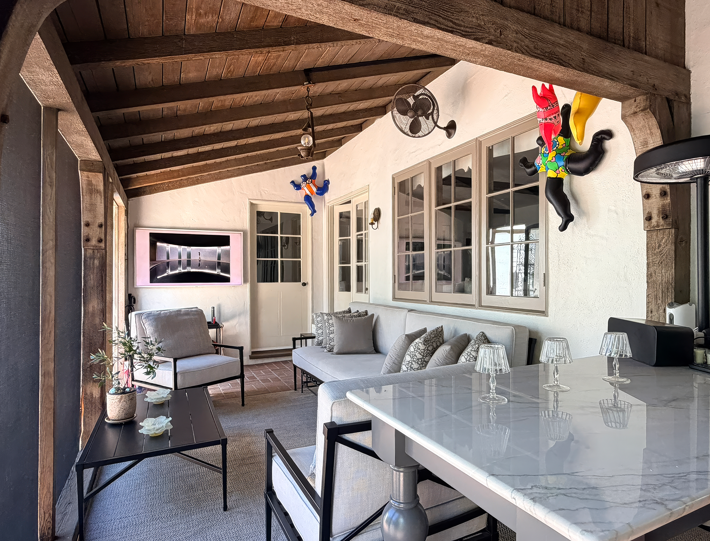 A cozy indoor space with a white wall, wooden ceiling beams, and decorative hanging figures of animals dressed in colorful clothes. There are multiple glass-paneled cabinets, a television, and a seating area with light-colored cushions. A white marbl