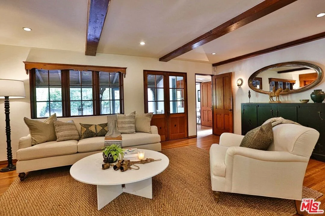 Living room with cream sofas, a round white coffee table, wooden floors, and large windows with wooden framing.