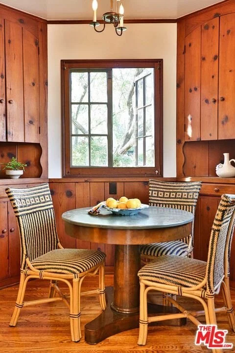 A cozy dining nook with a round wooden table surrounded by four rattan chairs with striped cushions. The area features wood-paneled walls and a window showing trees outside. A small plant and decorative vases are on the built-in wood cabinets, and a 