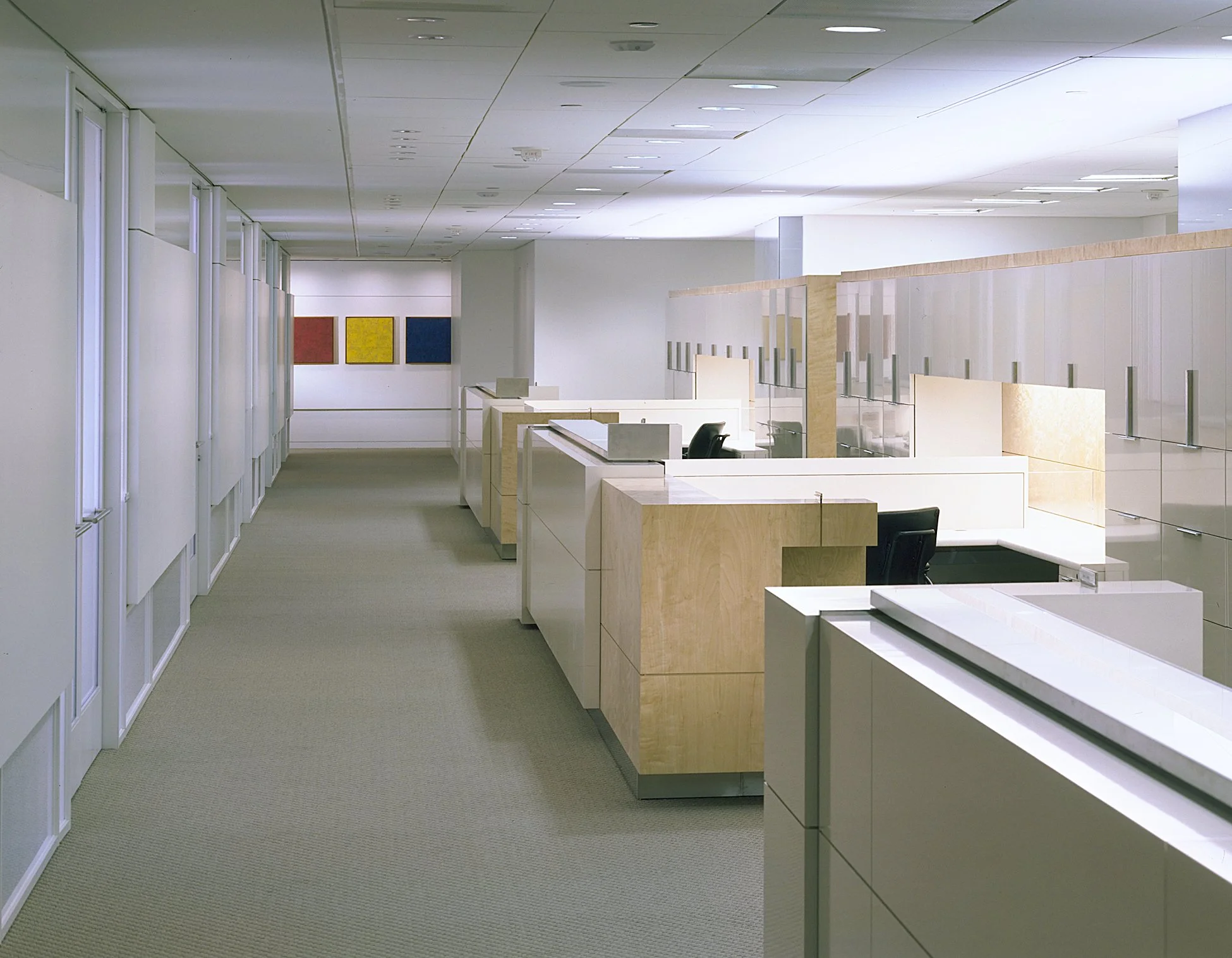 An empty modern office space with white walls, cubicles with wooden and glass partitions, and colorful artwork on the far wall.
